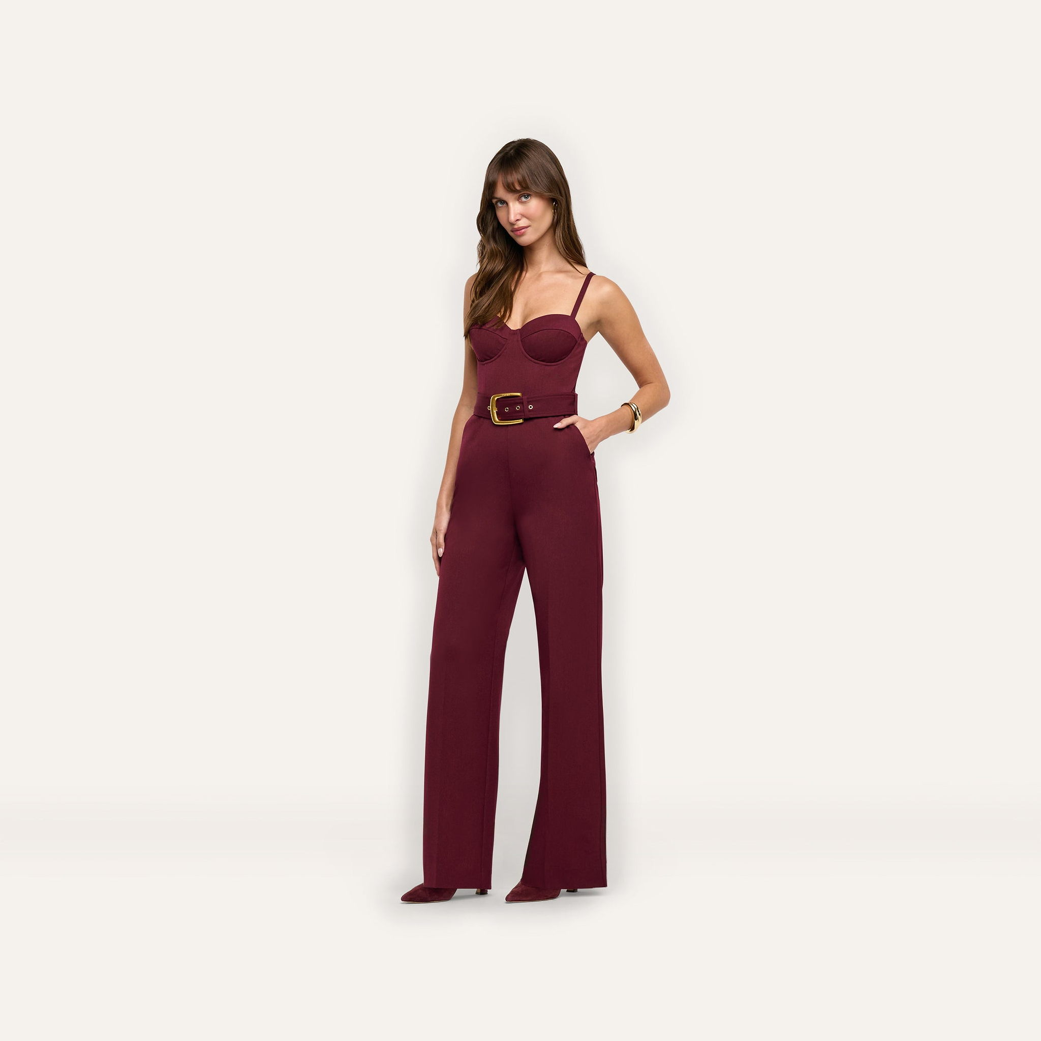 A woman wears the burgundy Jessa Bustier Jumpsuit, a sleeveless piece featuring a wide belt and straight-leg trousers.