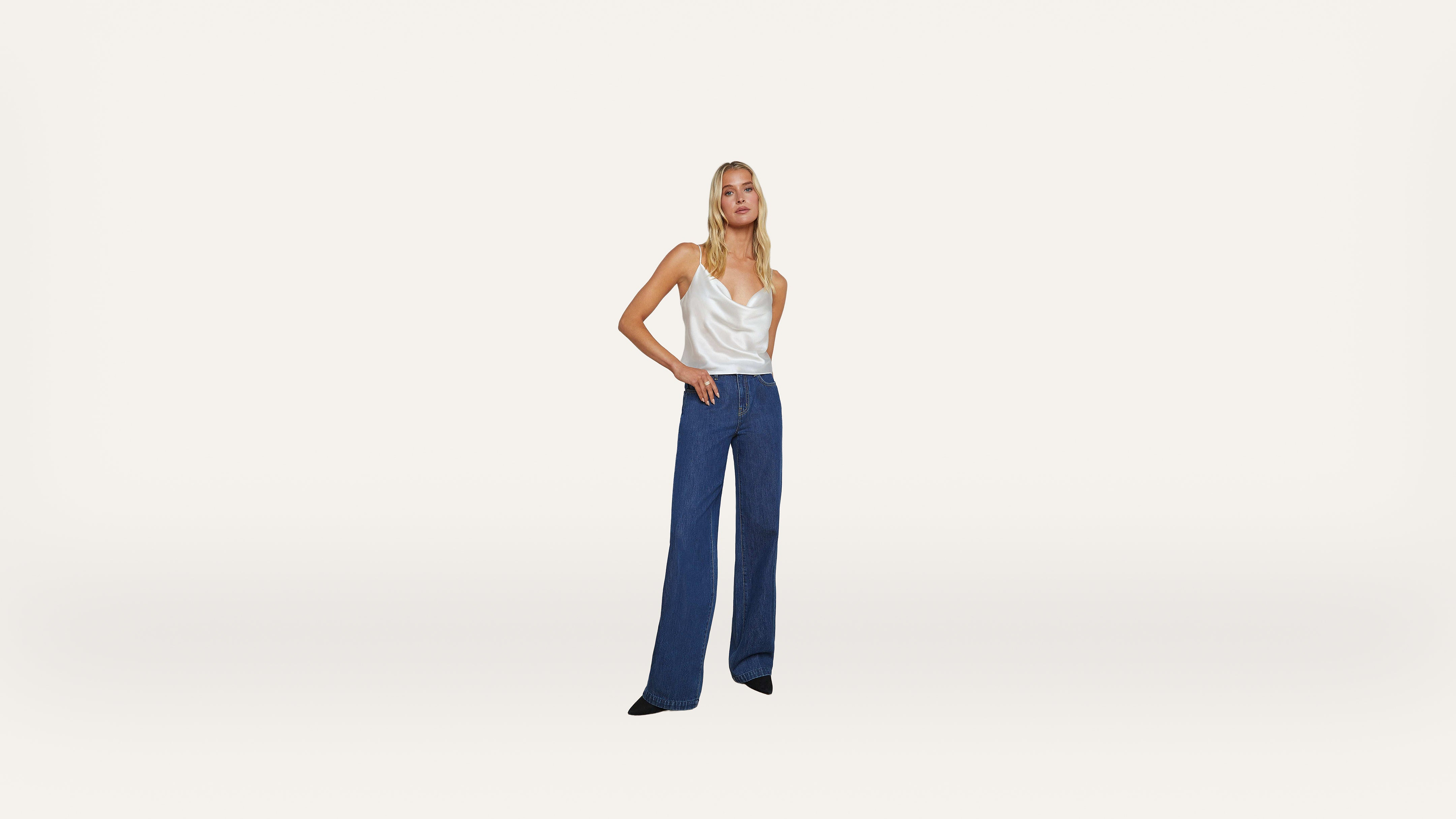 A woman stands against a plain white background, wearing a white sleeveless top and high-waisted, wide-leg blue jeans. Her left hand is in her pocket while her right arm rests at her side.