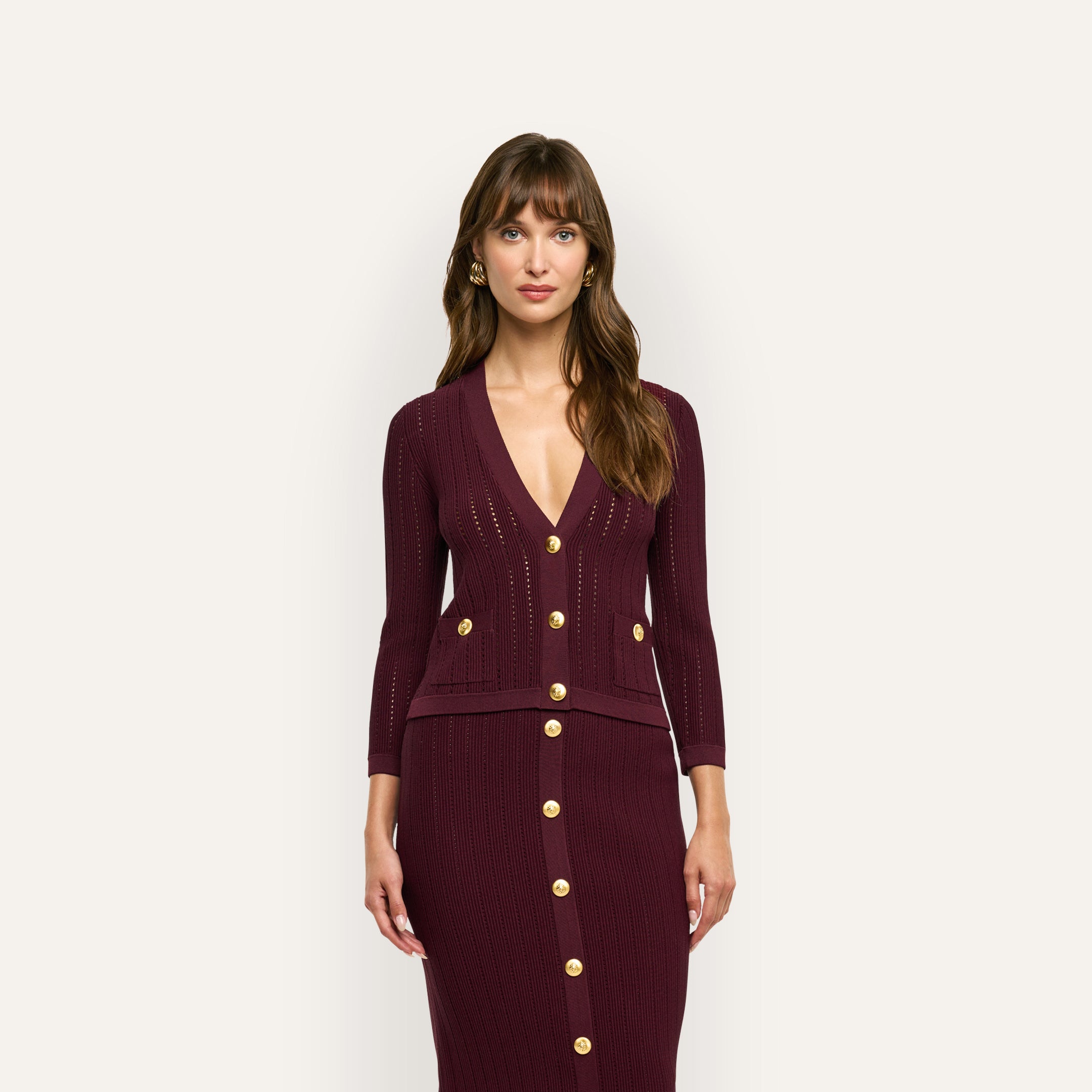 A model wears a deep red, pointelle-knit Irvin Pointelle Knit Cardigan with gold buttons and a matching button-front skirt, serving as a lightweight transitional layer.