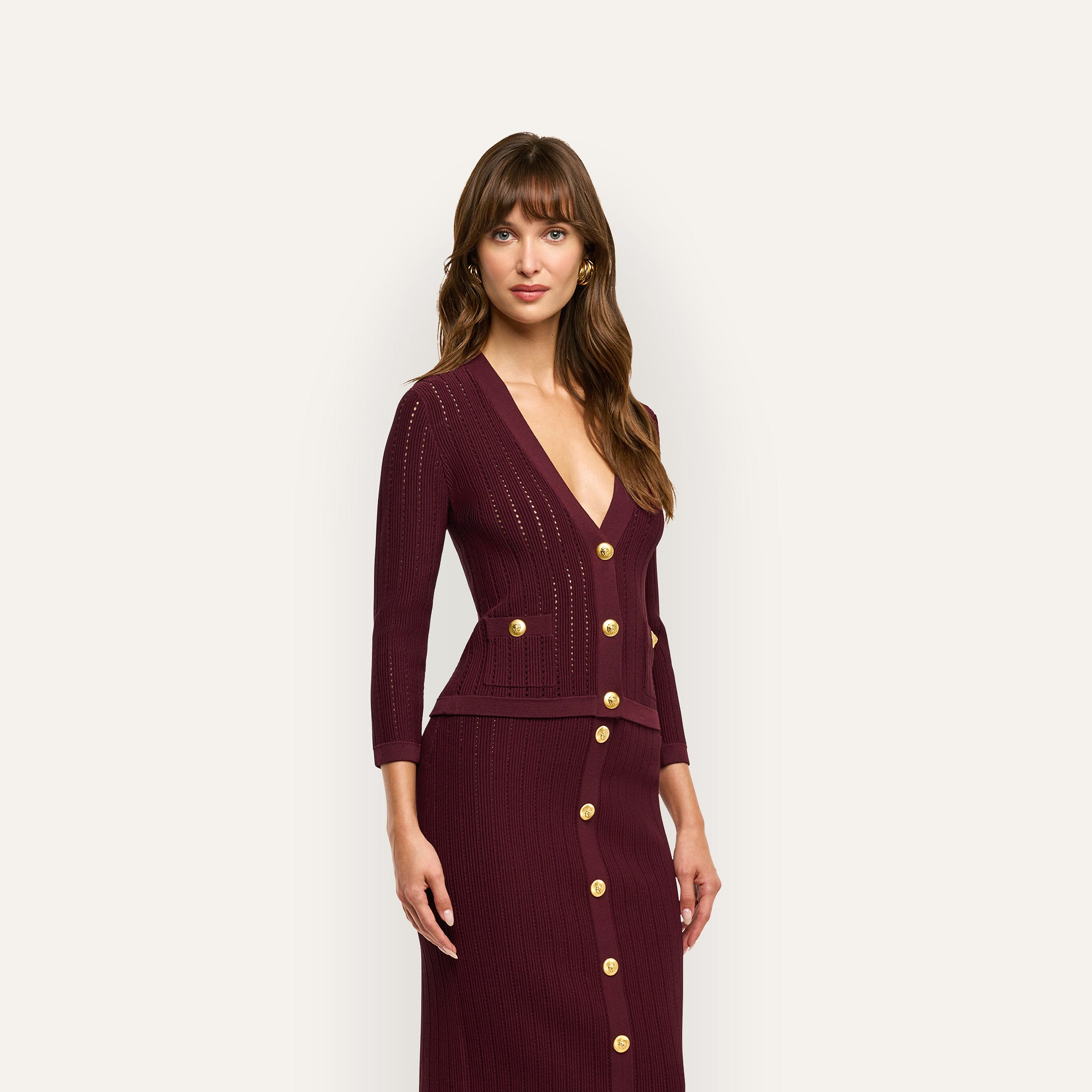 A woman wears a deep burgundy Irvin Pointelle Knit Cardigan with gold buttons over a matching ribbed midi skirt, serving as a lightweight transitional layer.