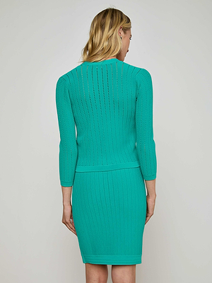 A blonde woman stands facing away from the camera in a fitted, textured turquoise Irvin Pointelle Knit Cardigan and matching skirt against a plain background.