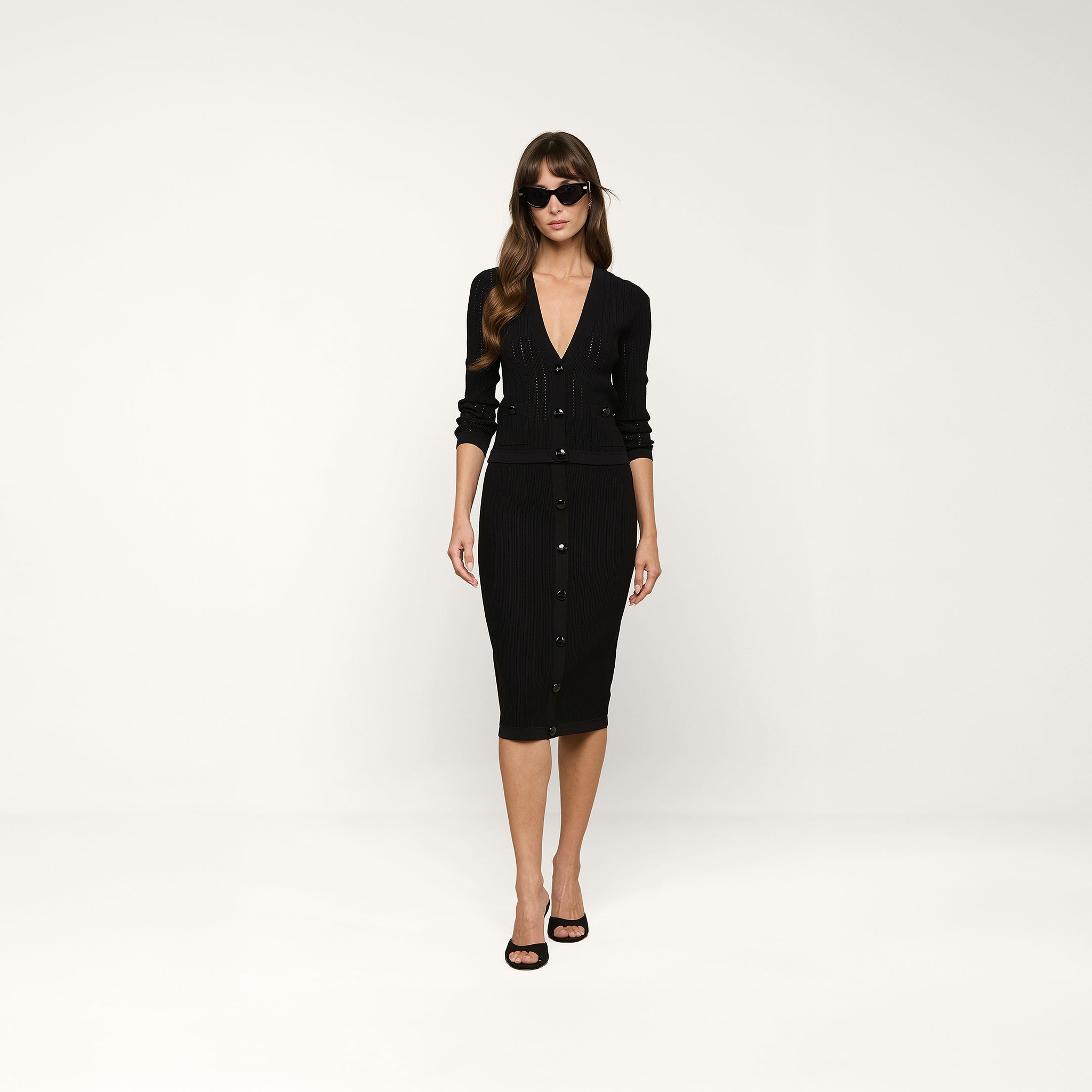 Woman wearing a fitted black button-down dress with long sleeves and black heels, standing against a plain white background. She has long hair, is wearing dark sunglasses, and completes her look with the Irvin Pointelle Knit Cardigan draped elegantly over her shoulders.