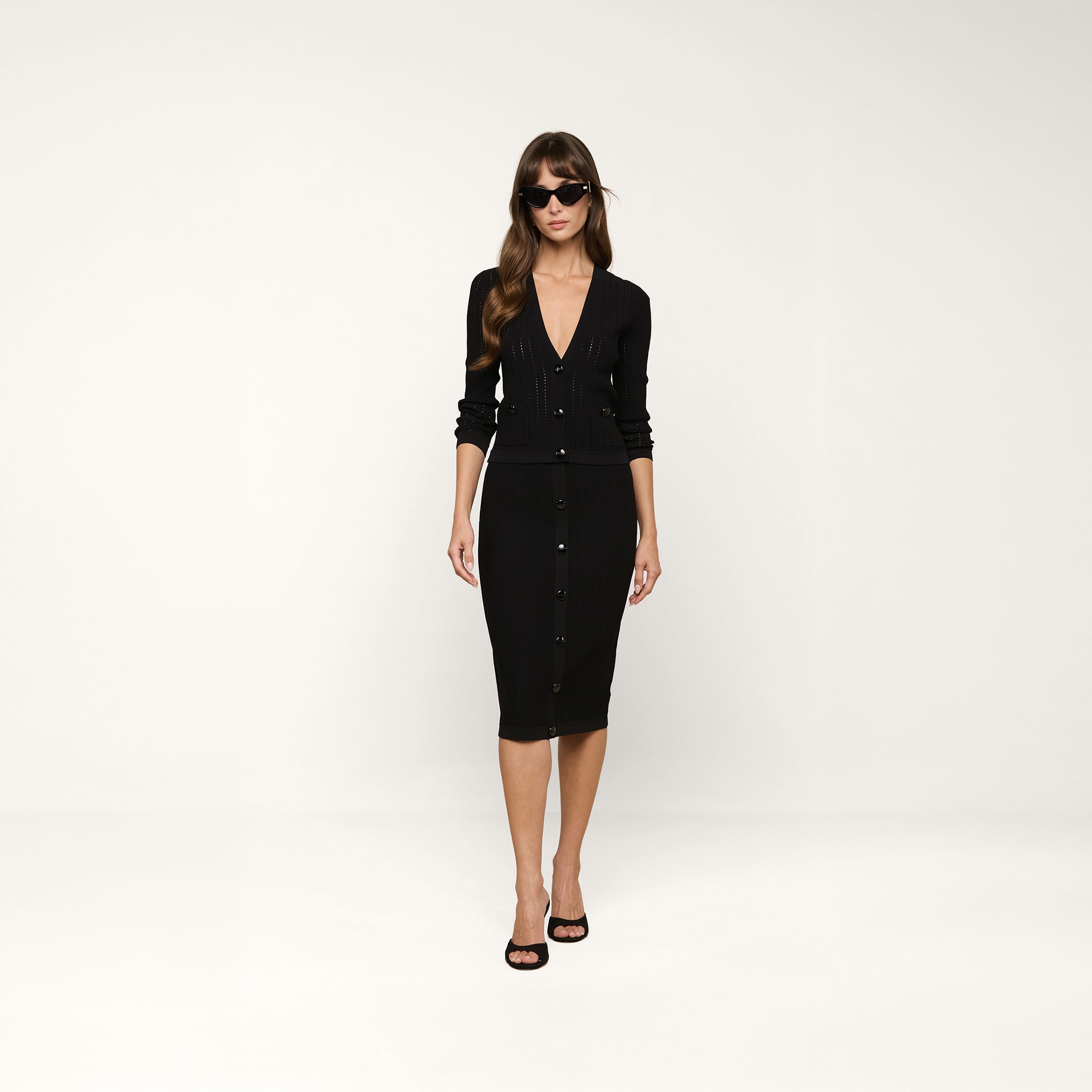 Woman wearing a fitted black button-down dress with long sleeves and black heels, standing against a plain white background. She has long hair, is wearing dark sunglasses, and completes her look with the Irvin Pointelle Knit Cardigan draped elegantly over her shoulders.