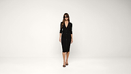 Woman in sunglasses wearing a black, button-down cardigan and matching black knee-length skirt with black high-heeled sandals stands against a plain white background.