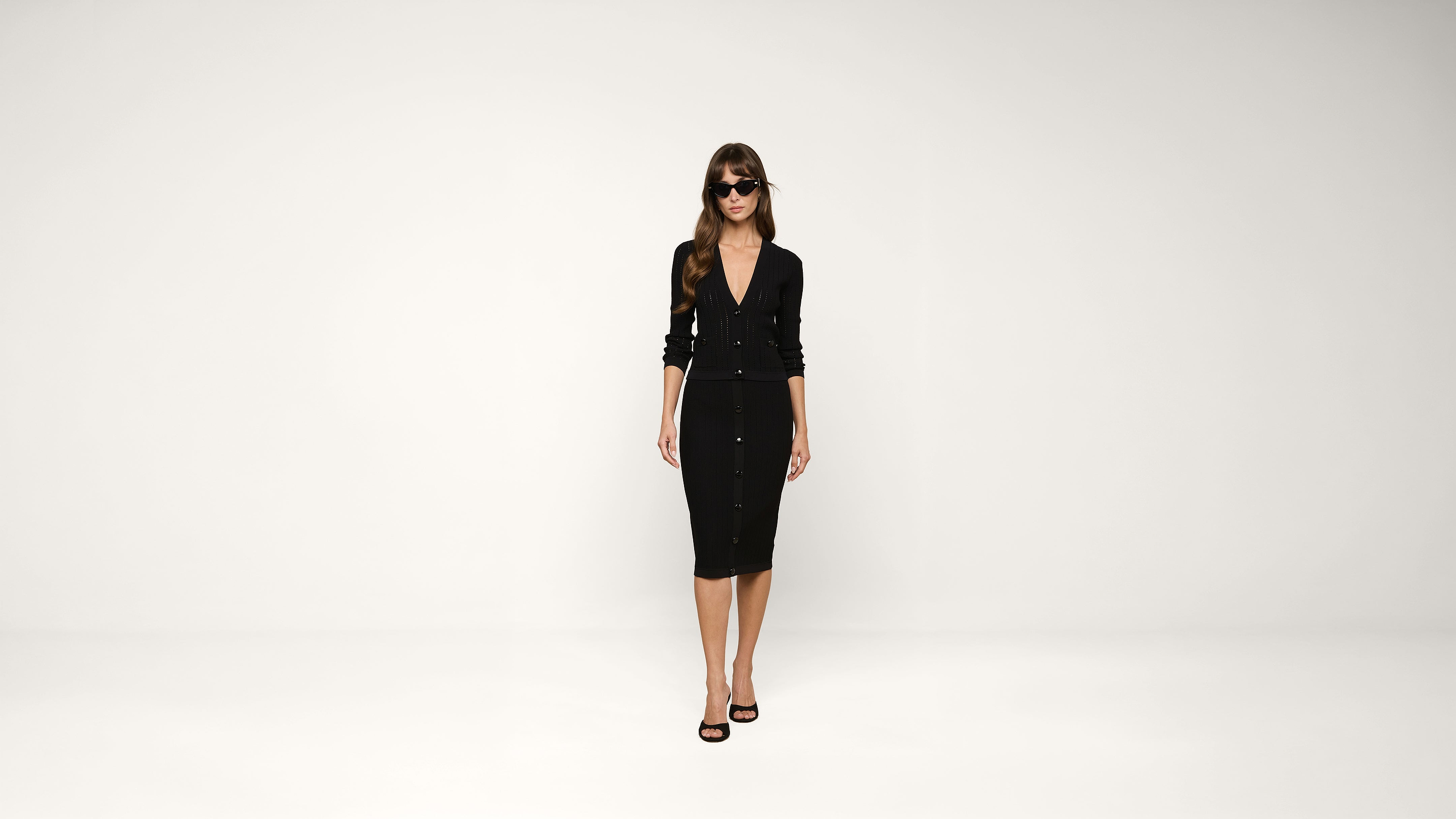 Woman in sunglasses wearing a black, button-down cardigan and matching black knee-length skirt with black high-heeled sandals stands against a plain white background.