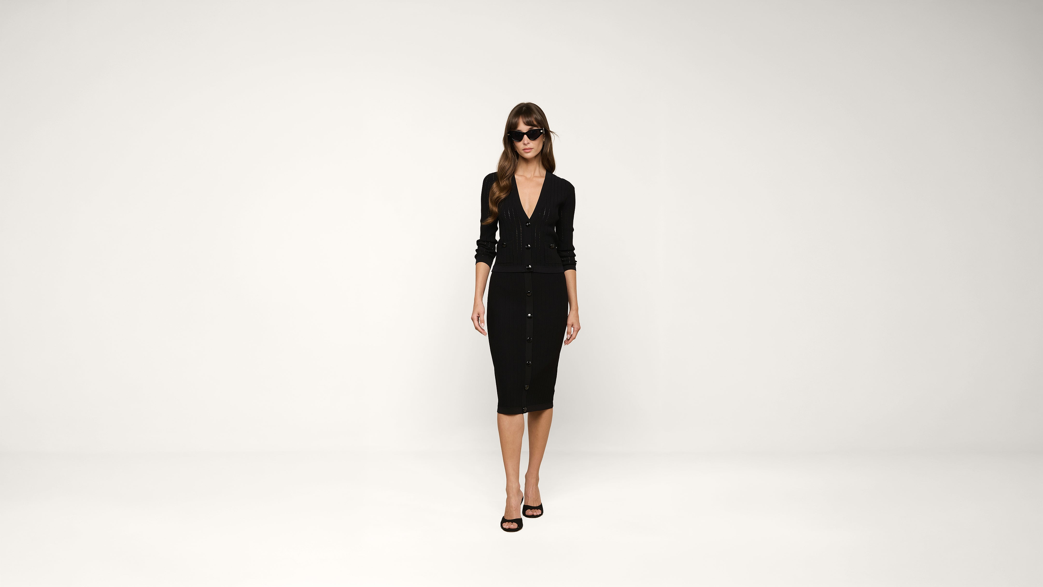 Woman in sunglasses wearing a black, button-down cardigan and matching black knee-length skirt with black high-heeled sandals stands against a plain white background.