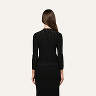 A woman with long brown hair, wearing a fitted black long-sleeve dress and the Irvin Pointelle Knit Cardigan, is standing facing away from the camera against a plain light background.