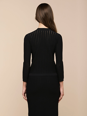 A woman with long brown hair faces away, wearing the Irvin Pointelle Knit Cardigan over a fitted black dress, set against a neutral beige background.