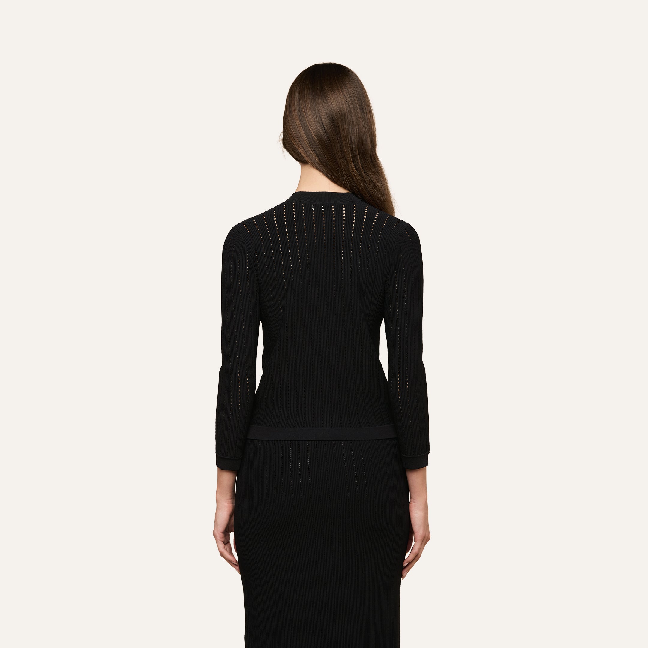 A woman with long brown hair, wearing a fitted black long-sleeve dress and the Irvin Pointelle Knit Cardigan, is standing facing away from the camera against a plain light background.