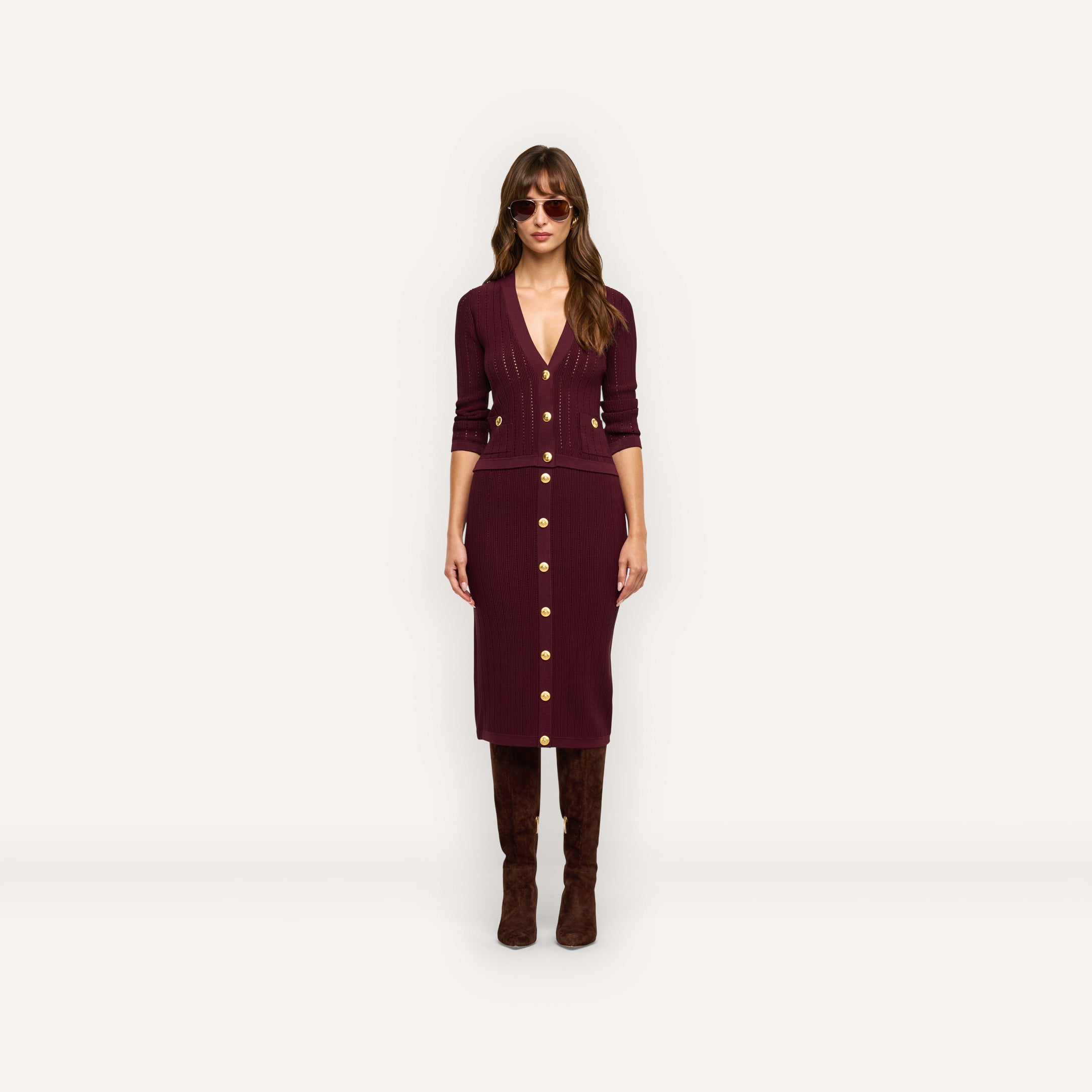 A woman wears a dark red, button-down Melania Pointelle Knit Skirt and a matching long-sleeved top with gold buttons, paired with brown knee-high boots and sunglasses.