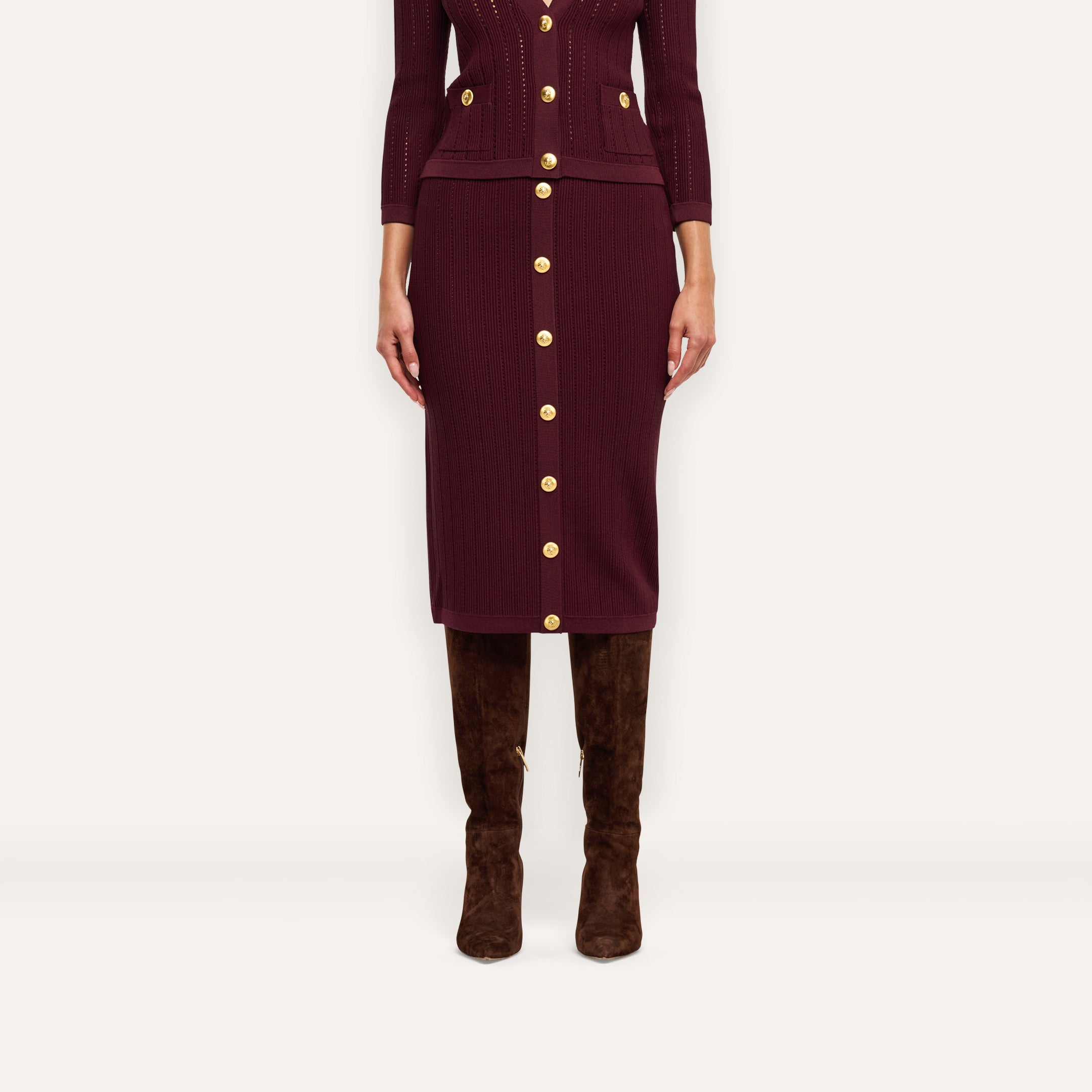 The Melania Pointelle Knit Skirt is a burgundy knit midi skirt with an elastic waistband and gold buttons down the center front, worn with brown over-the-knee boots.