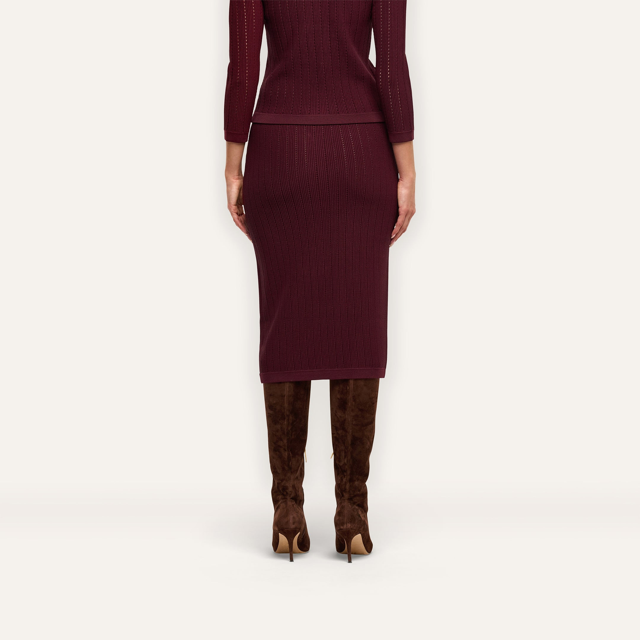 A person seen from behind wears a maroon Melania Pointelle Knit Skirt with gold buttons and brown knee-high heeled boots.