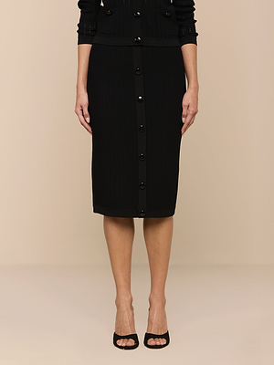 A person wears the Melania Pointelle Knit Skirt—a body-skimming, black, knee-length style—with a matching black top and open-toe heels, standing against a plain beige background.