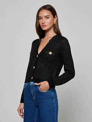 A woman wears the L'AGENCE Blanca Sequin Cardigan and blue high-waisted jeans against a light gray background. She has long brown hair and gold hoop earrings.
