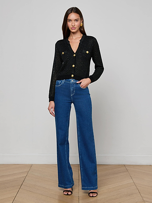 A woman with long brown hair poses indoors against a light gray wall, wearing the L'AGENCE Blanca Sequin Cardigan, high-waisted blue jeans, black heeled sandals, and gold earrings.