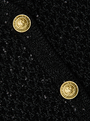 Close-up of the L'AGENCE Blanca Sequin Cardigan in textured black fabric with two shiny embossed dome buttons featuring an eagle and wreath, sewn diagonally for added detail to this sophisticated lightweight layer.