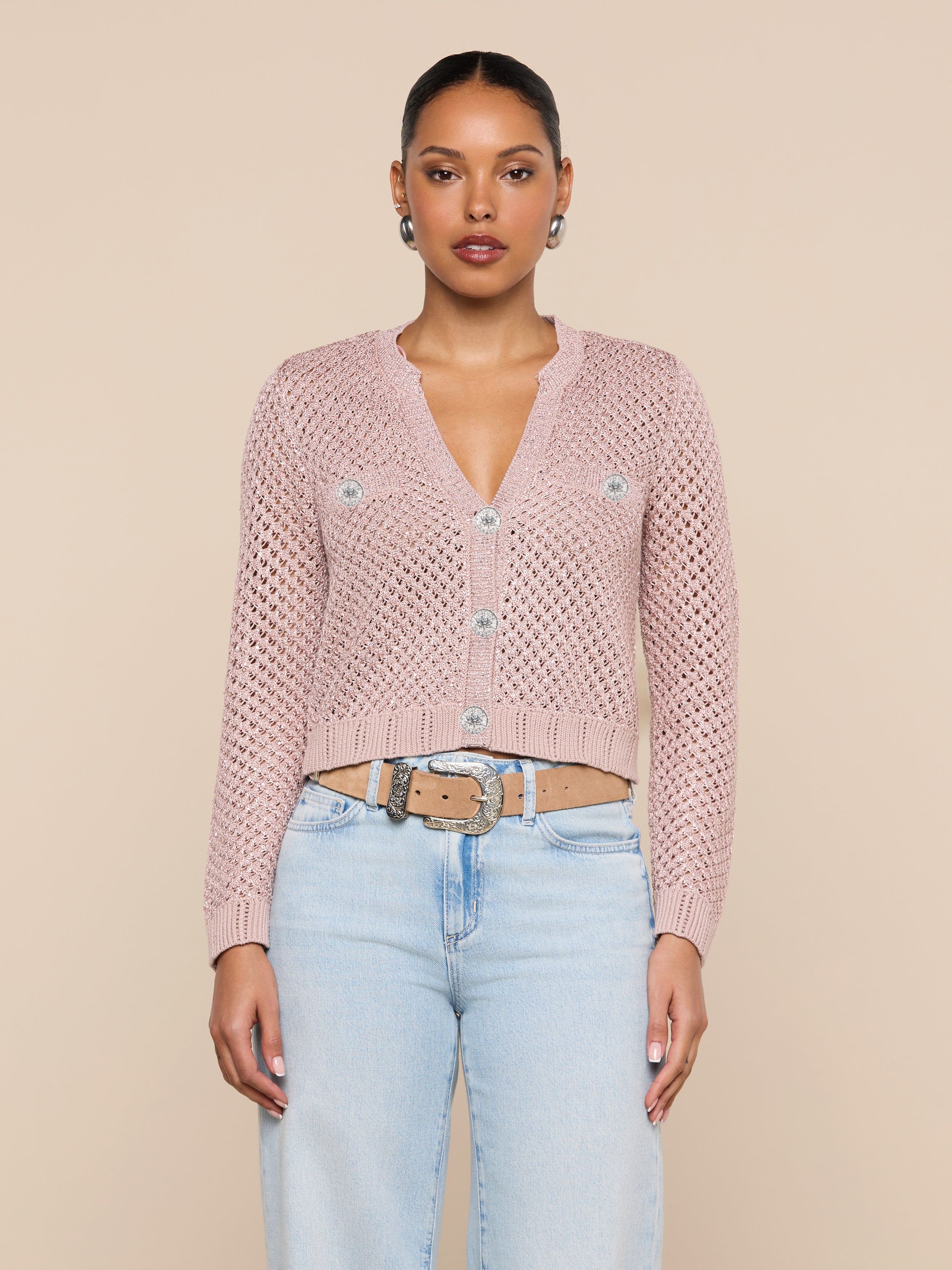 A woman stands facing forward, wearing the Blanca Sequin Cardigan in adobe rose pink as a lightweight layer, paired with light blue jeans, a beige belt, and silver stud earrings. Her hair is pulled back, and she poses against a plain off-white background.