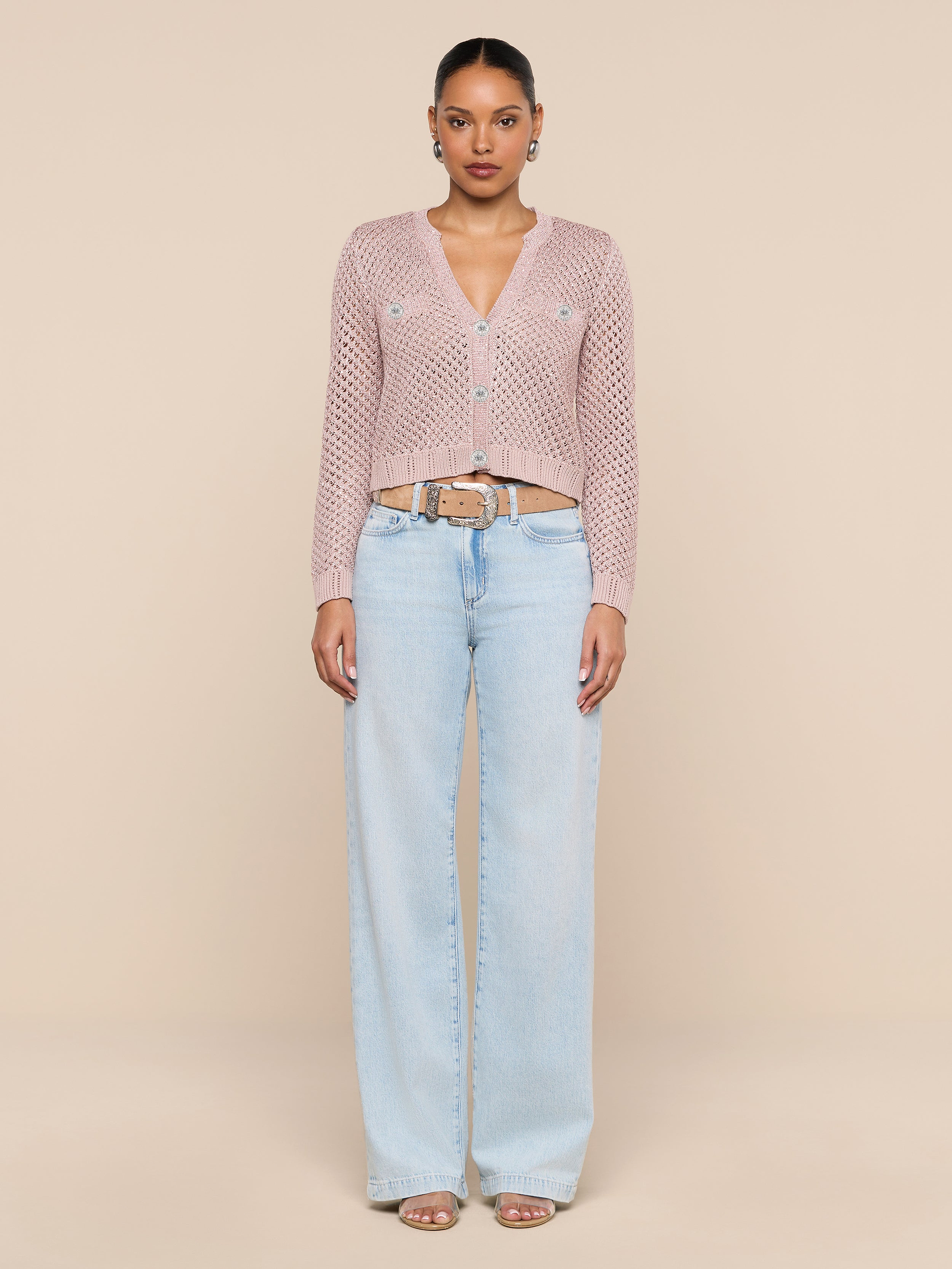 A woman stands against a plain white background, wearing the Blanca Sequin Cardigan in adobe rose, paired with light blue wide-leg jeans, a beige belt, hoop earrings, and open-toed sandals.