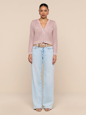 A person stands facing forward against a beige background, wearing the Blanca Sequin Cardigan, light blue high-waisted wide-leg jeans, a tan belt, and open-toe heels.