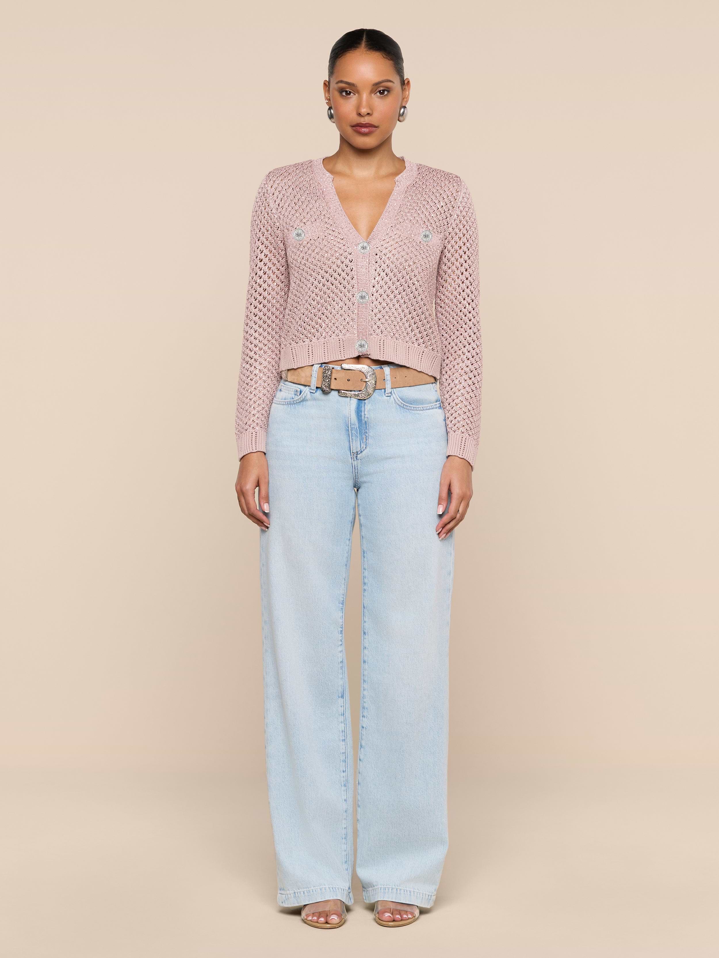 A woman stands against a plain white background, wearing the Blanca Sequin Cardigan in adobe rose, paired with light blue wide-leg jeans, a beige belt, hoop earrings, and open-toed sandals.