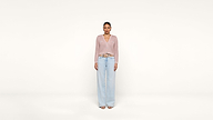 A woman stands facing forward against a plain white background, wearing a light pink knit cardigan, light blue wide-leg jeans, and open-toe sandals.