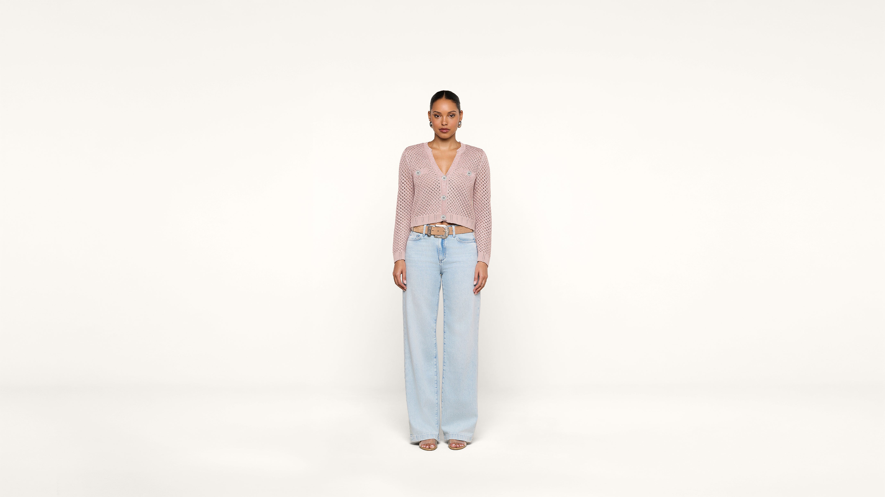 A woman stands facing forward against a plain white background, wearing a light pink knit cardigan, light blue wide-leg jeans, and open-toe sandals.