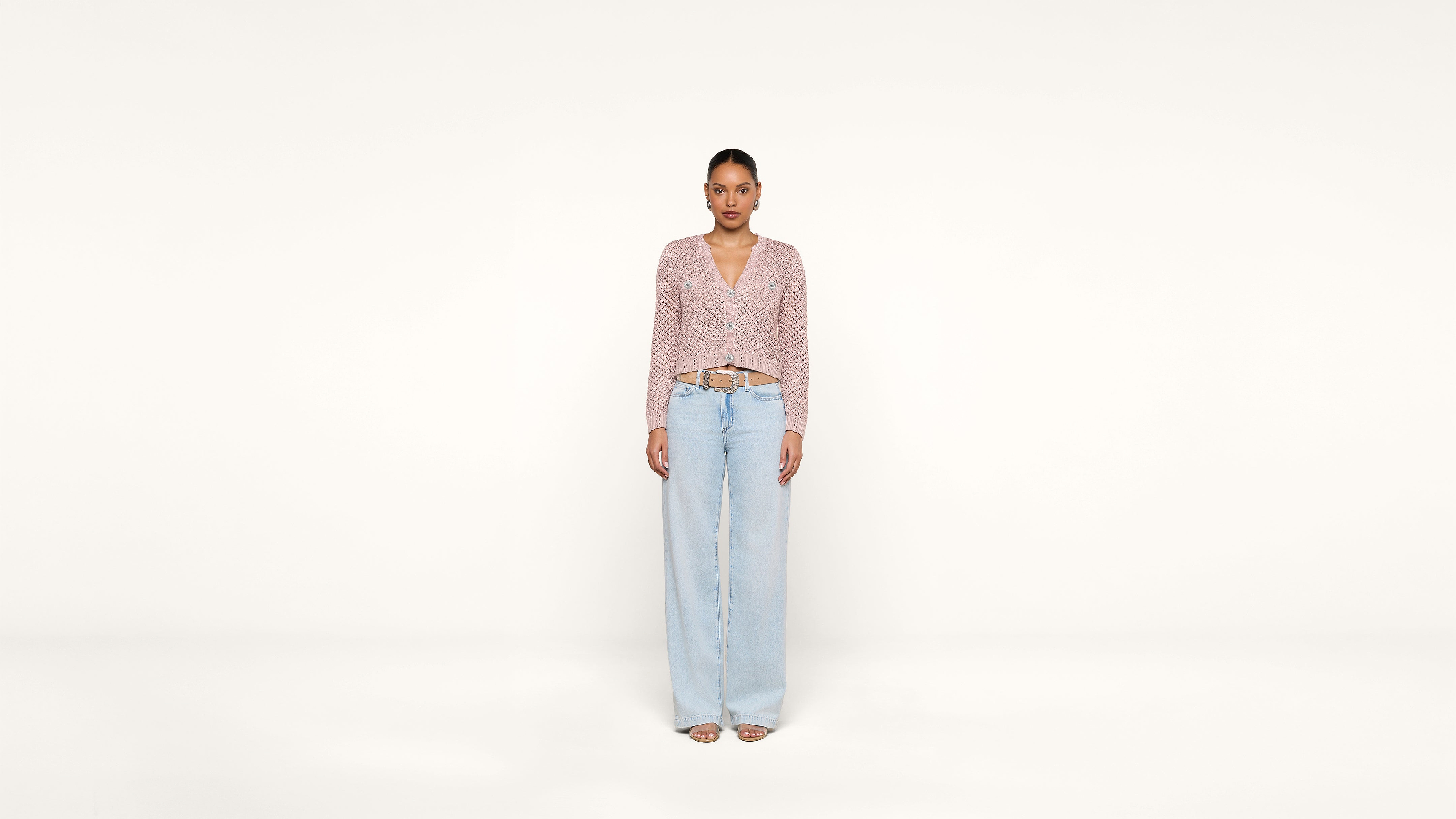 A woman stands facing forward against a plain white background, wearing a light pink knit cardigan, light blue wide-leg jeans, and open-toe sandals.