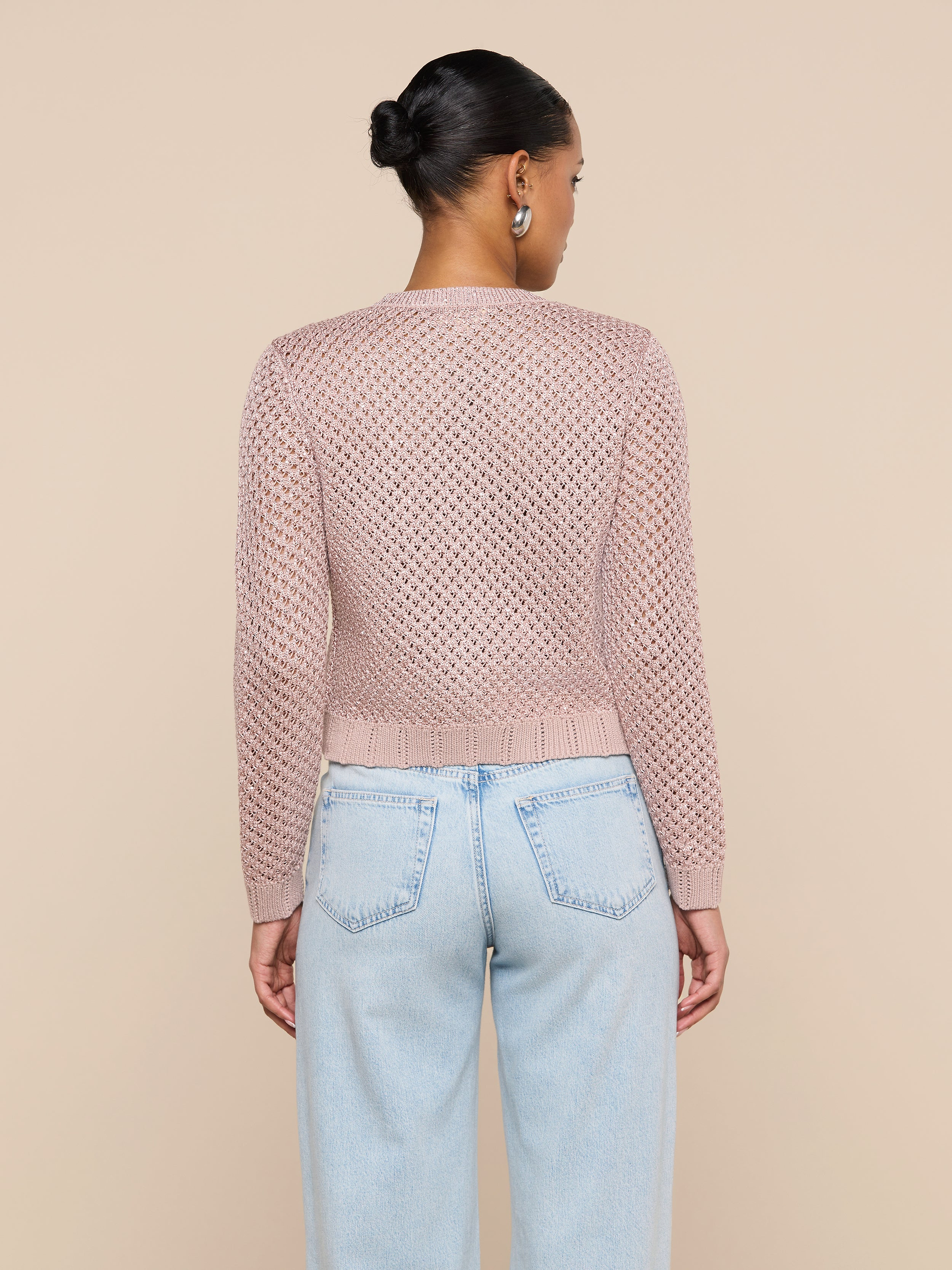 A person stands facing away, wearing the Blanca Sequin Cardigan in adobe rose paired with light blue jeans. Their hair is styled in a neat bun, set against a plain, light background.