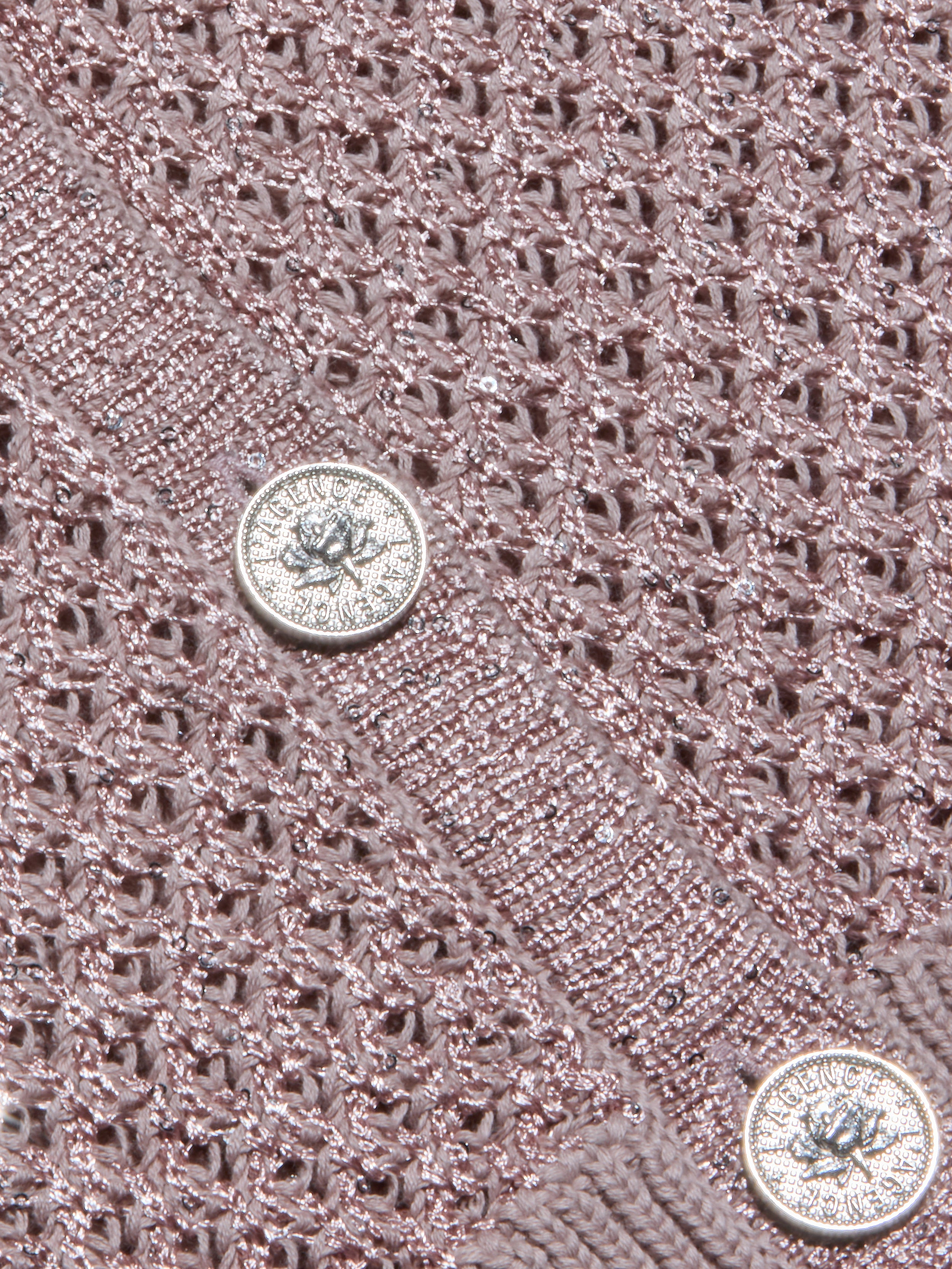 Close-up of the Blanca Sequin Cardigan’s textured adobe rose knit, featuring two shiny silver buttons with embossed designs and text. This lightweight layer displays a lacy, open pattern, with the buttons sewn onto a solid knit band.