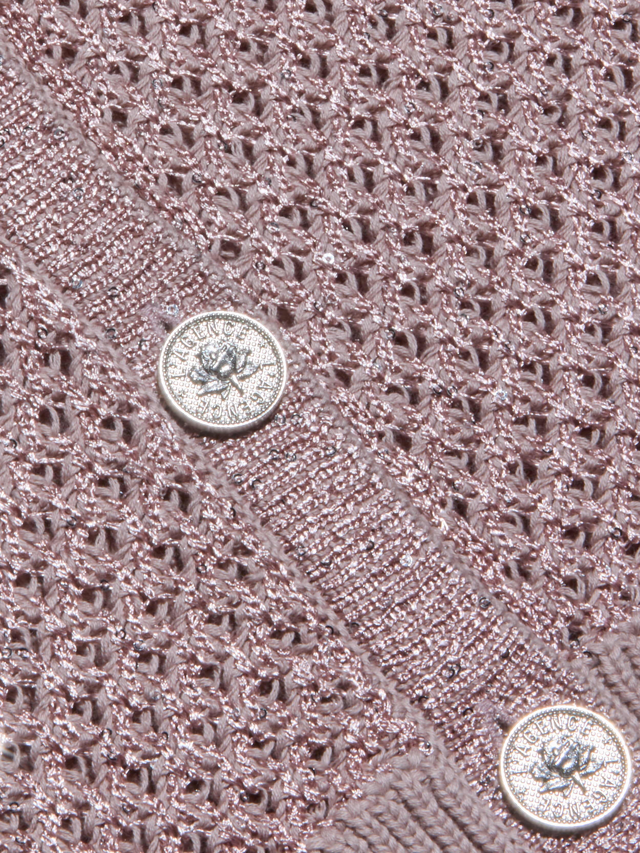 Close-up of the Blanca Sequin Cardigan’s textured adobe rose knit, featuring two shiny silver buttons with embossed designs and text. This lightweight layer displays a lacy, open pattern, with the buttons sewn onto a solid knit band.