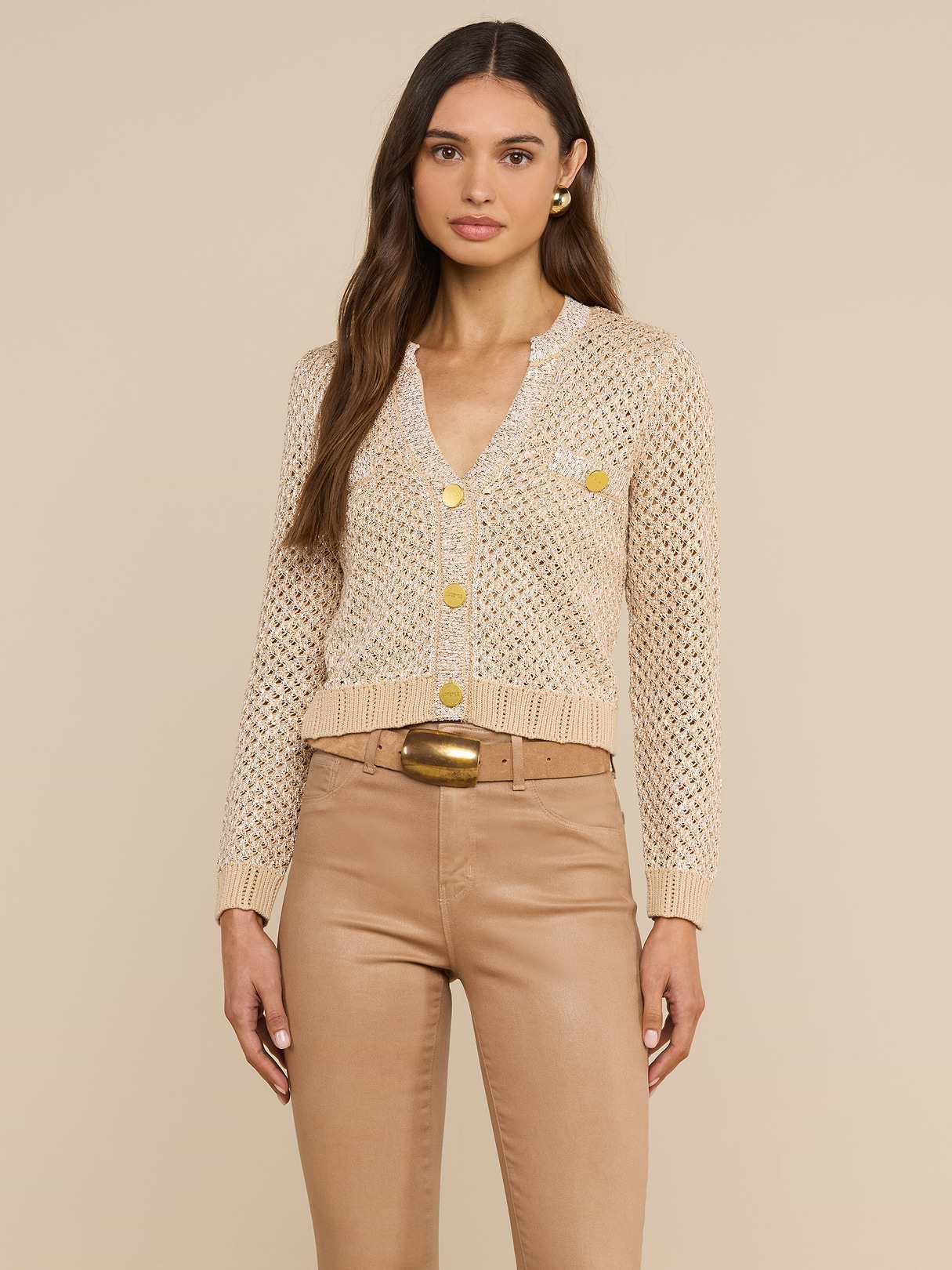 Une femme aux longs cheveux châtains porte le cardigan « Blanca Sequin » — un modèle en maille pointelle beige neutre orné de boutons dorés —, associé à un pantalon beige à taille haute et à une ceinture à boucle dorée, debout devant un fond beige uni.
