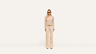 A woman wearing sunglasses, a beige long-sleeve top, wide-leg pants, and brown pointed shoes stands confidently against a plain white background.