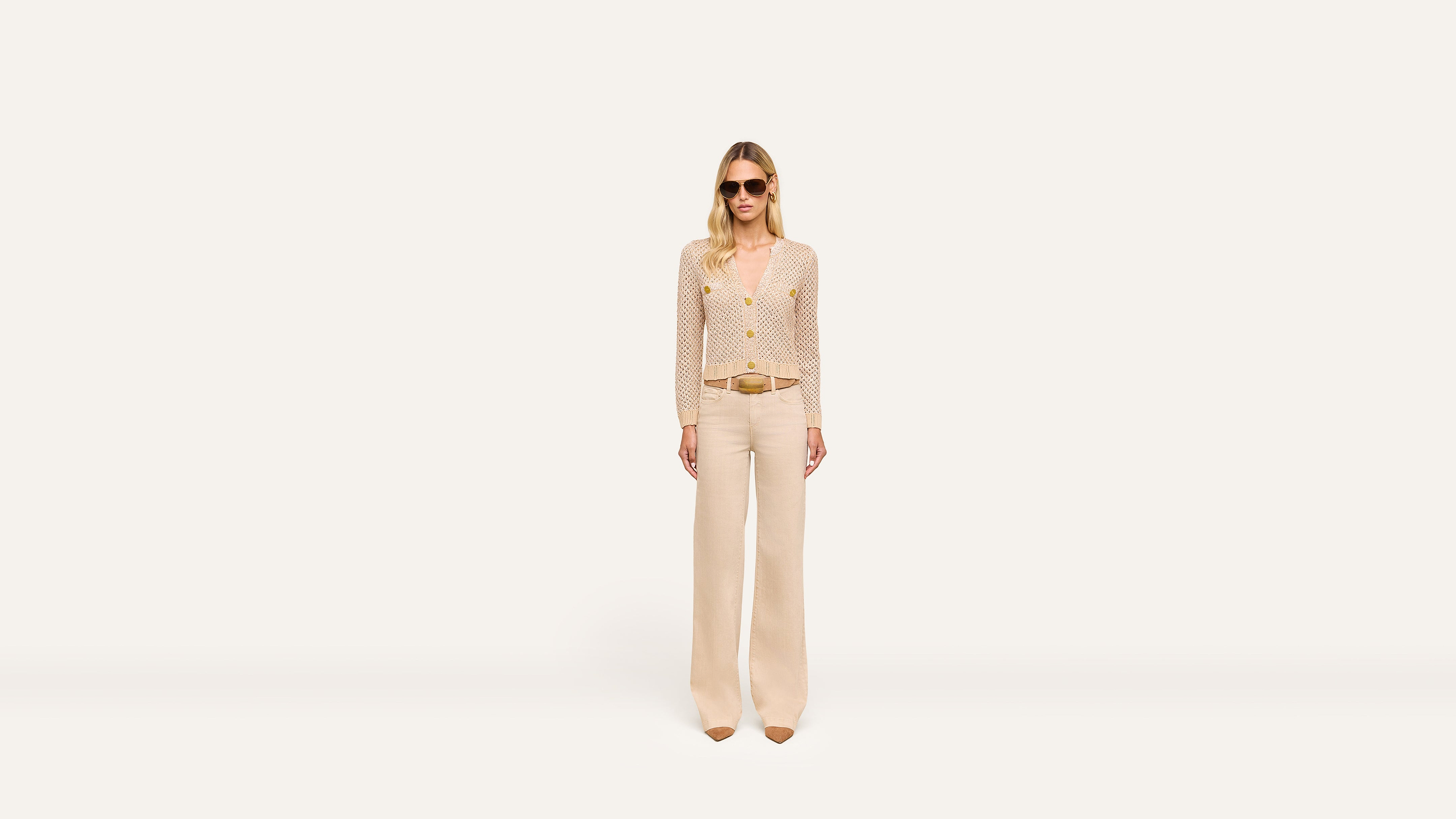 A woman wearing sunglasses, a beige long-sleeve top, wide-leg pants, and brown pointed shoes stands confidently against a plain white background.