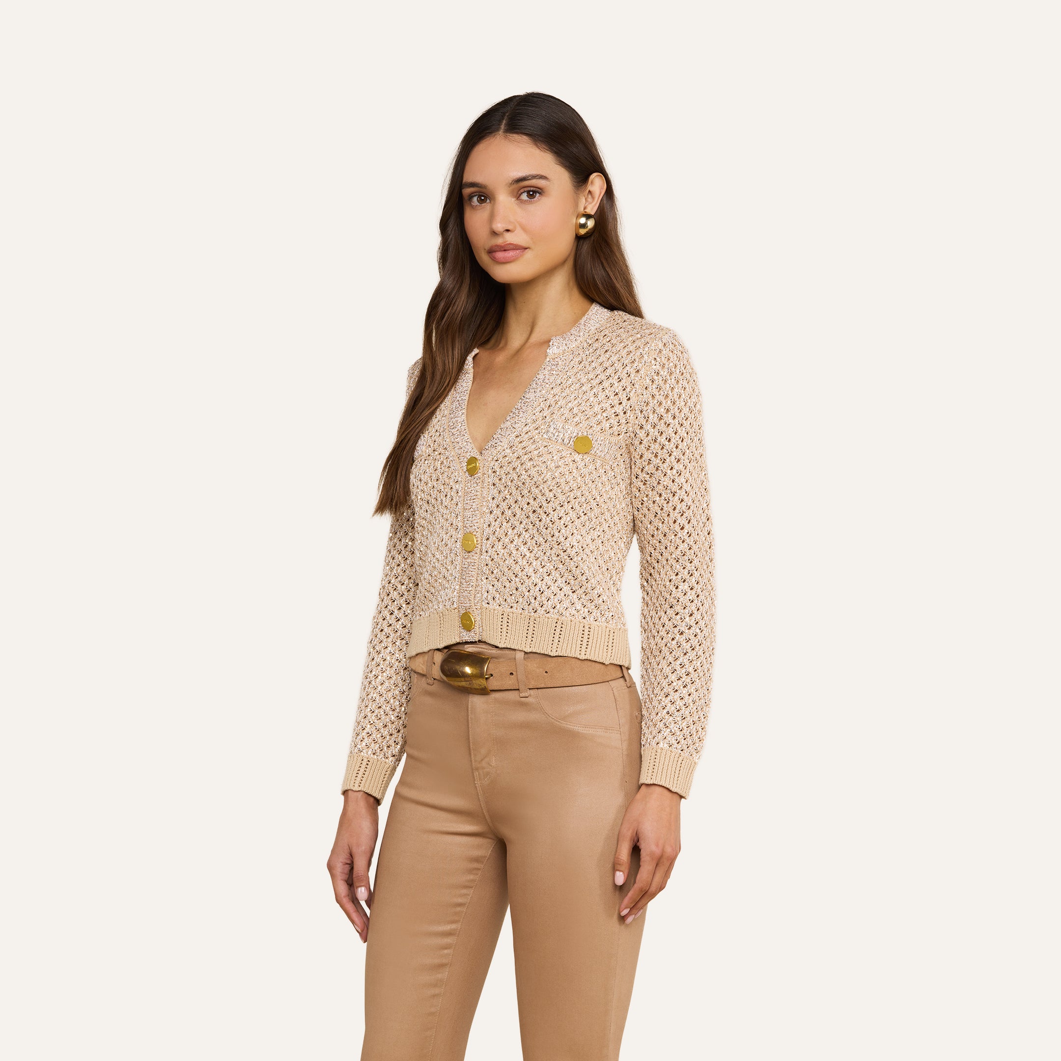 Une femme porte le cardigan « Blanca Sequin », un modèle en maille couleur beige neutre fermé par des boutons dorés, quelle a associé à un pantalon marron clair et à une large ceinture marron, debout devant un fond beige uni.