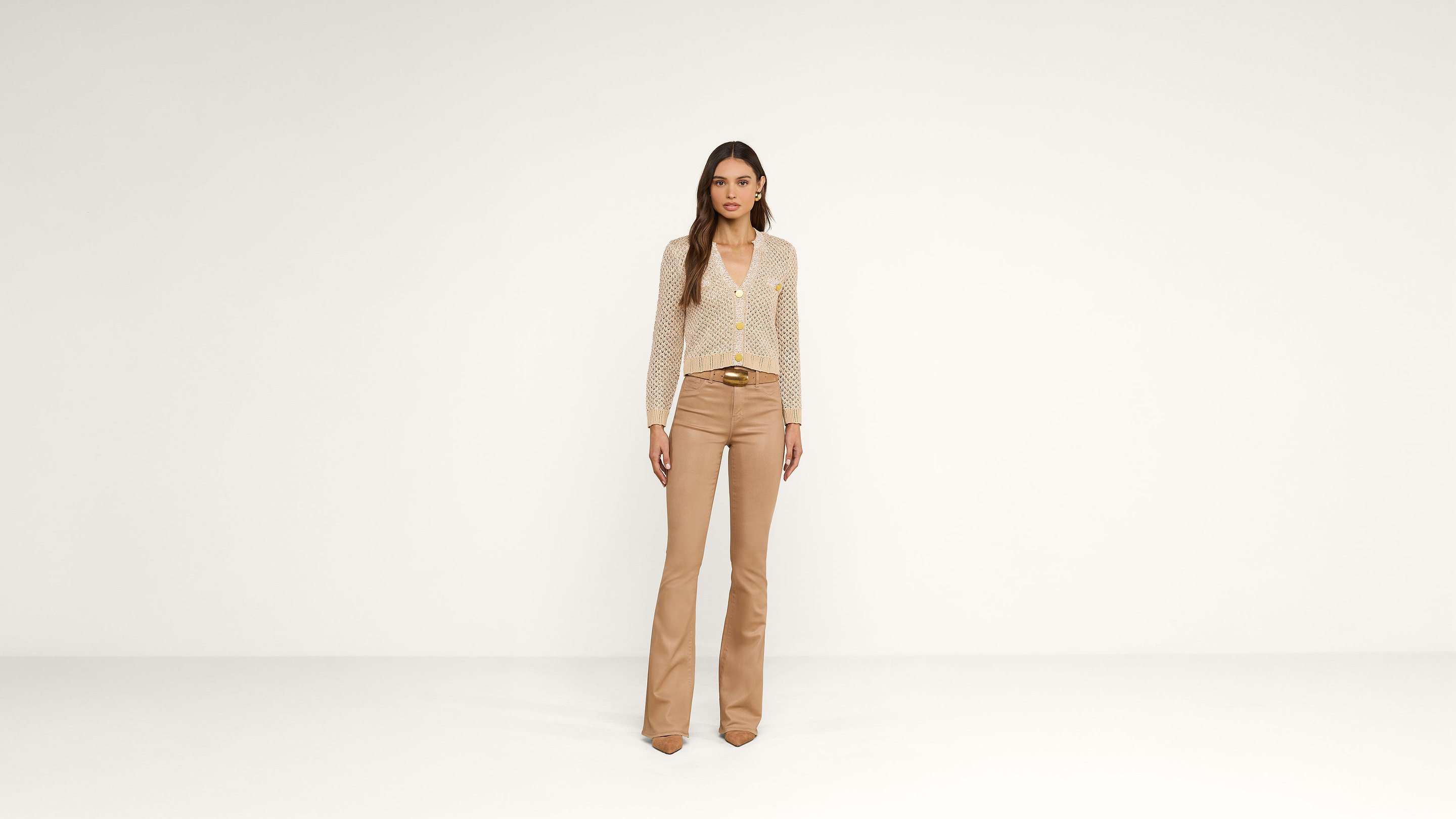 A woman stands against a plain white background wearing a light polka dot blouse, beige flared pants, and brown pointed shoes.