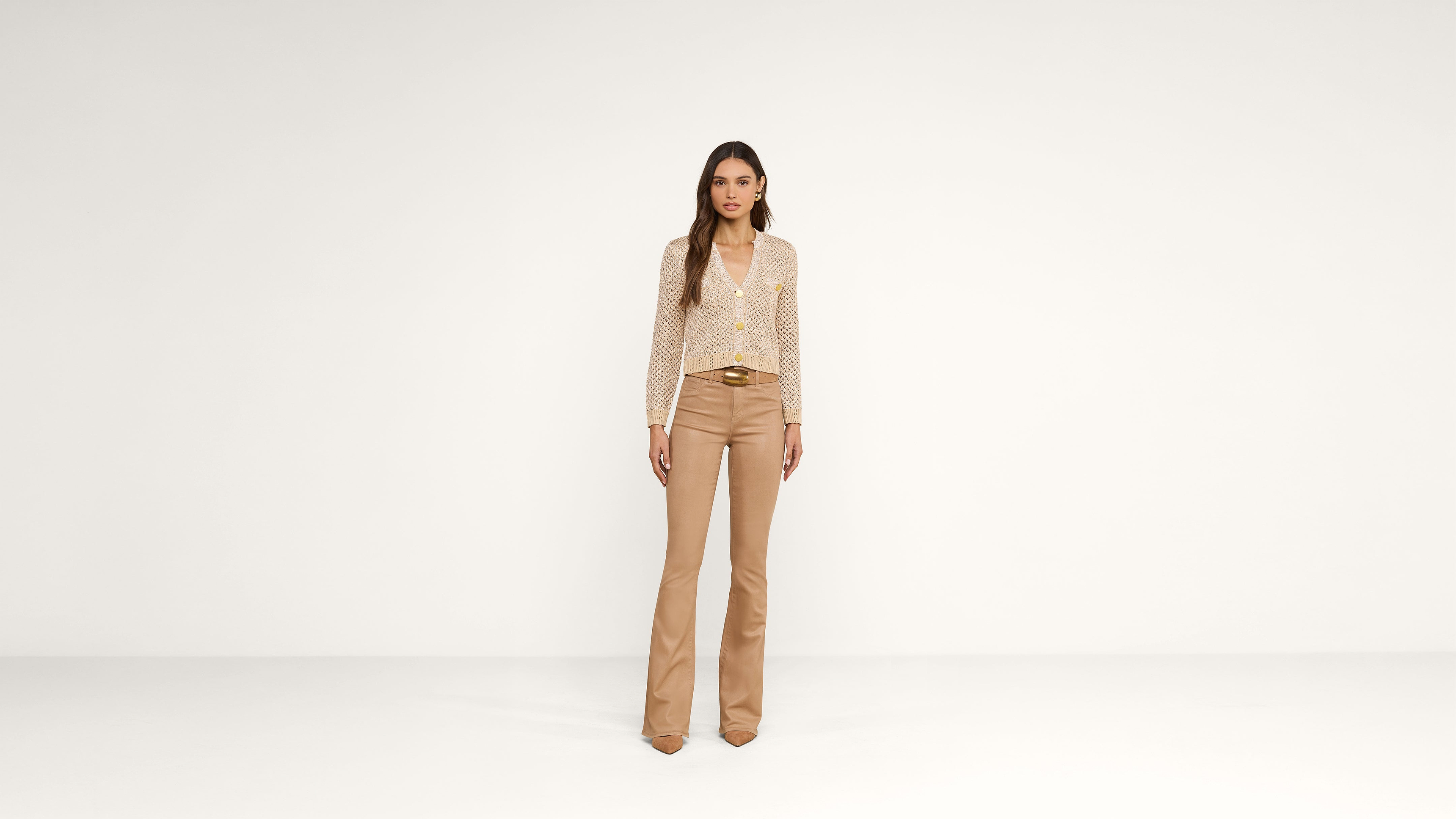 A woman stands against a plain white background wearing a light polka dot blouse, beige flared pants, and brown pointed shoes.