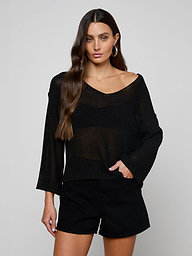 A woman with long brown hair wears the Ethan Knit Pullover—sheer black, off-shoulder—and black shorts, standing against a plain light gray background with one hand in her pocket, looking at the camera.