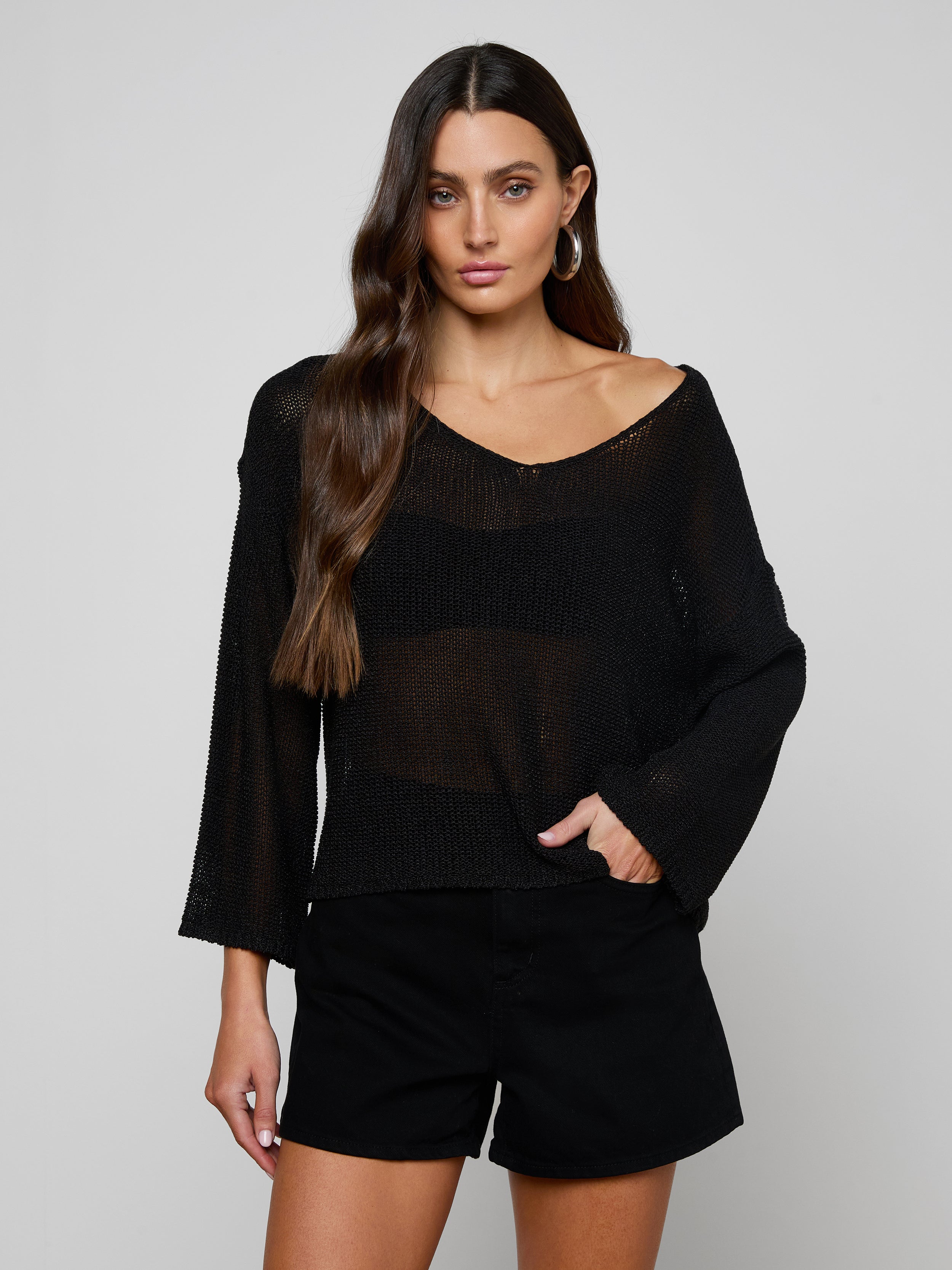 A woman with long brown hair wears the Ethan Knit Pullover—sheer black, off-shoulder—and black shorts, standing against a plain light gray background with one hand in her pocket, looking at the camera.