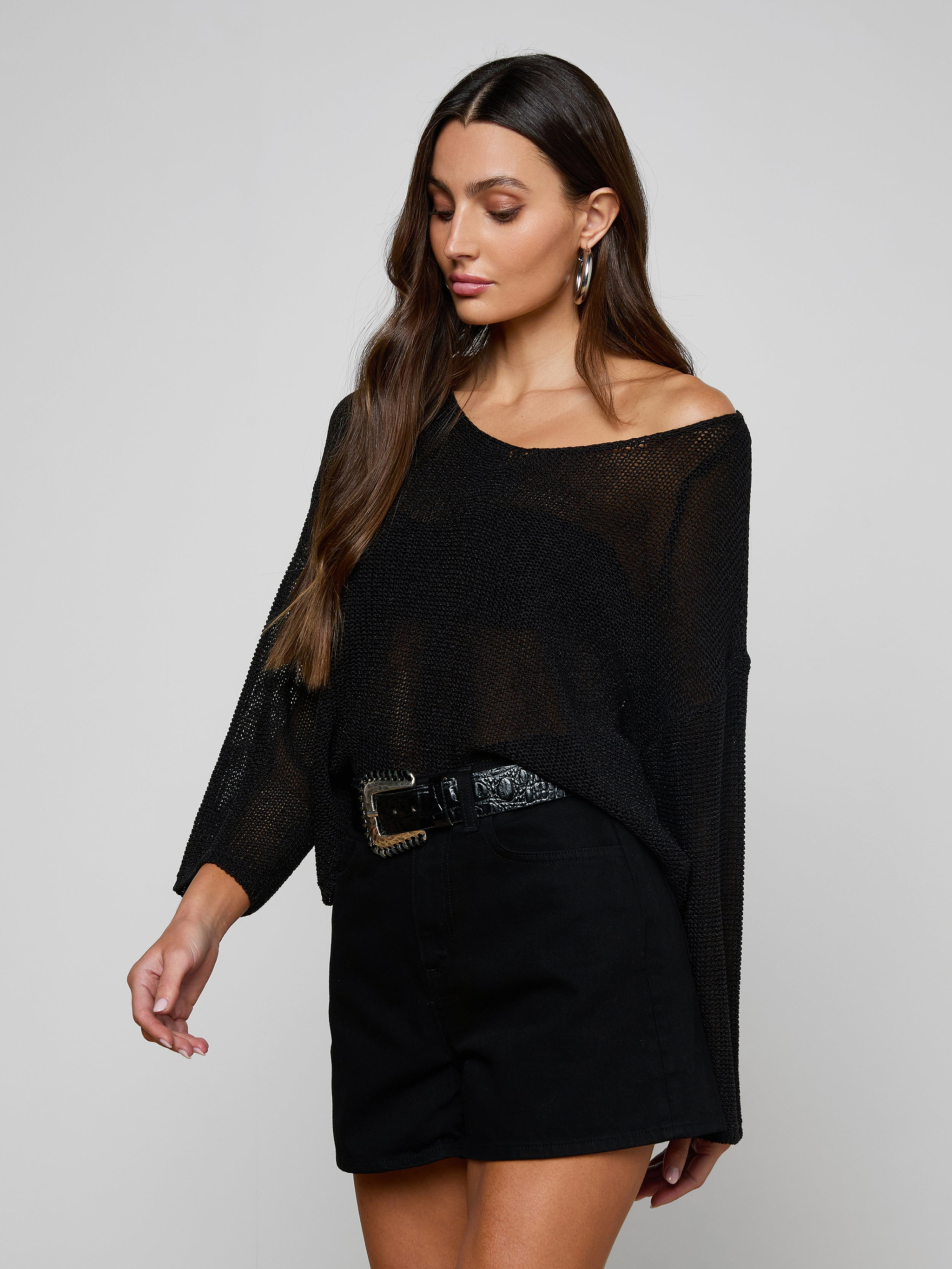 A woman with long brown hair wears the Ethan Knit Pullover, a sheer black knit top with a relaxed fit, paired with black high-waisted shorts and a wide belt. She looks down and slightly to the side against a plain background.