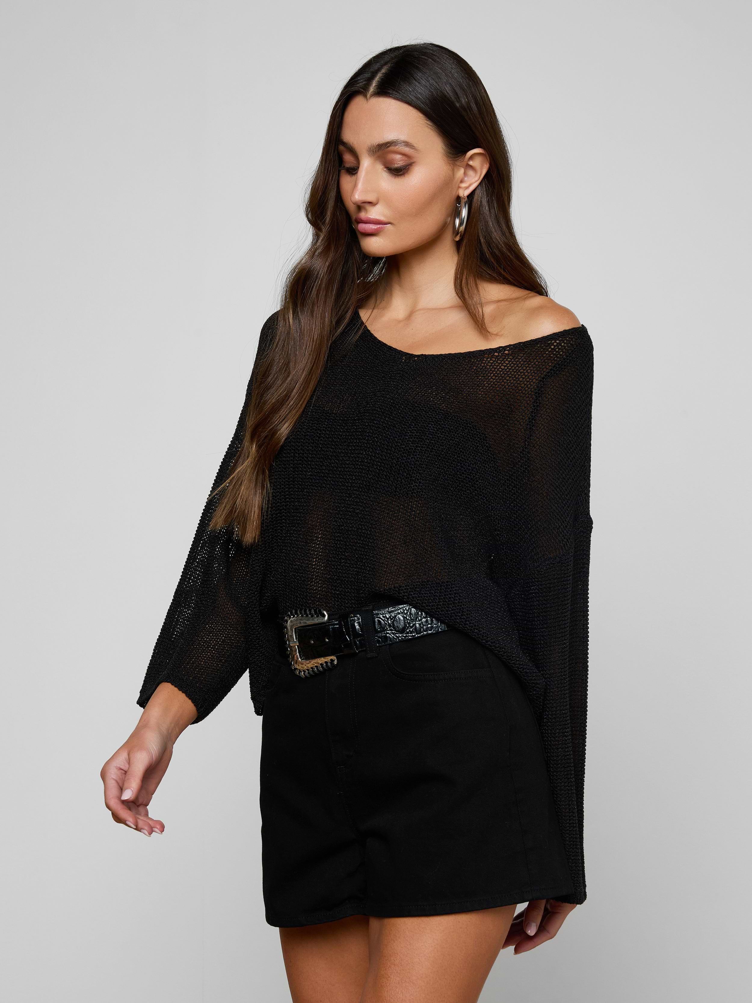 A woman with long brown hair wears the Ethan Knit Pullover, a sheer black knit top with a relaxed fit, paired with black high-waisted shorts and a wide belt. She looks down and slightly to the side against a plain background.