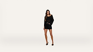 A woman stands against a plain white background wearing a sheer black top, black shorts, and black heeled sandals. She has long, wavy hair and a relaxed pose with one hand on her hip.