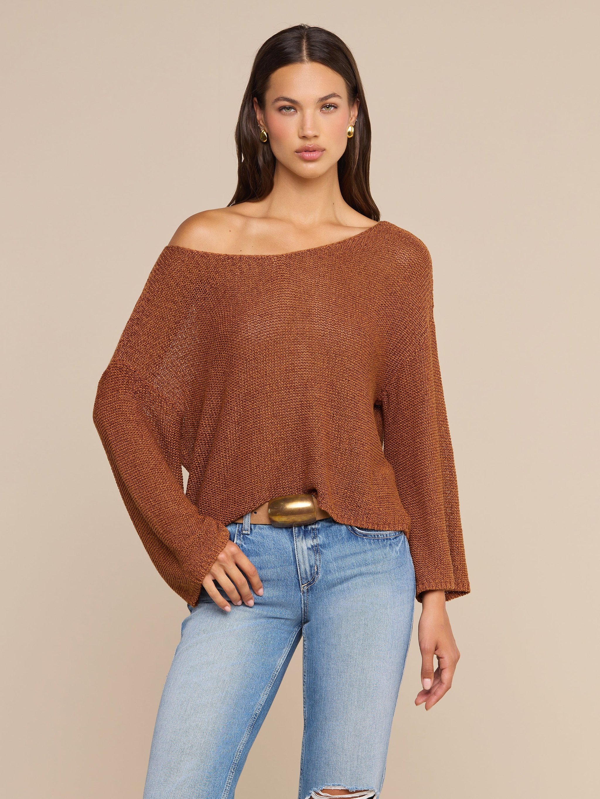 한 여성이 앞을 향해 서 있는데, 넉넉한 핏의 갈색 ‘Ethan Knit Pullover’ 오프숄더 상의에 하늘색 찢어진 청바지와 금색 벨트를 매치하고 있다. 그녀는 긴 갈색 머리에 금색 귀걸이를 하고 있으며, 베이지색 배경 앞에 서 있다.