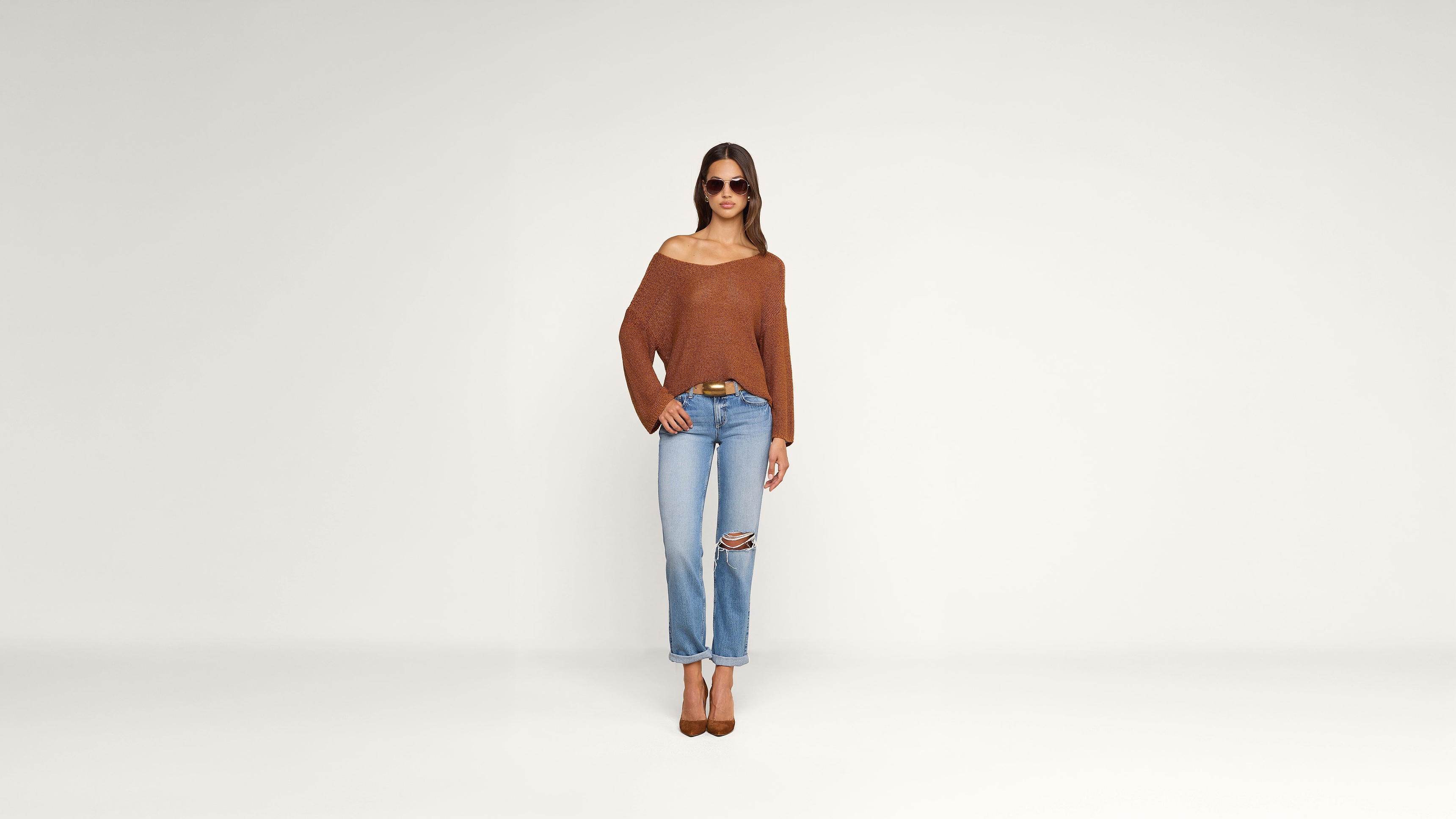 Woman wearing sunglasses, a loose brown sweater, ripped light blue jeans, and brown heels stands against a plain white background, looking straight ahead.