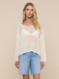 A woman stands facing forward against a plain light background, wearing the Ethan Knit Pullover—a sheer, cream, long-sleeve top with a relaxed fit—layered over a white bralette and paired with knee-length denim shorts.