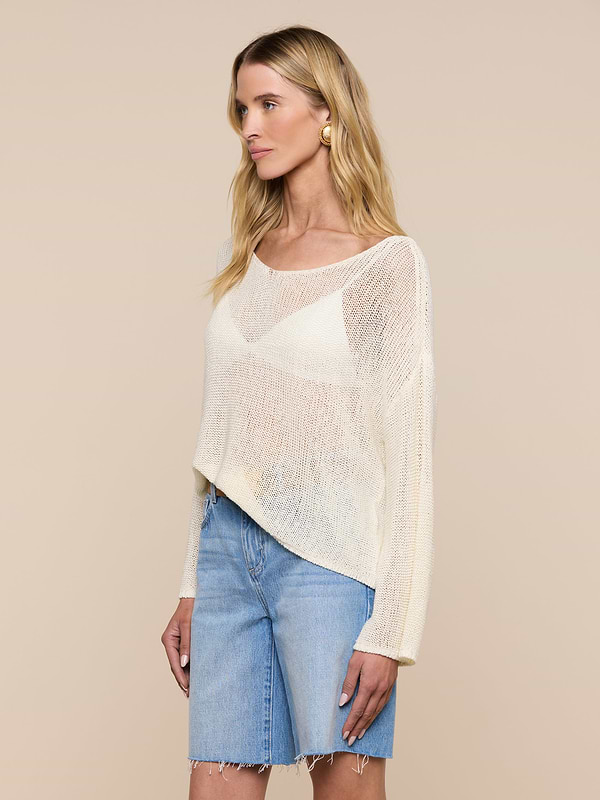 A woman with long blonde hair wears the Ethan Knit Pullover in a relaxed fit with an off-shoulder neckline over a white top and blue knee-length denim shorts, standing against a plain light background.