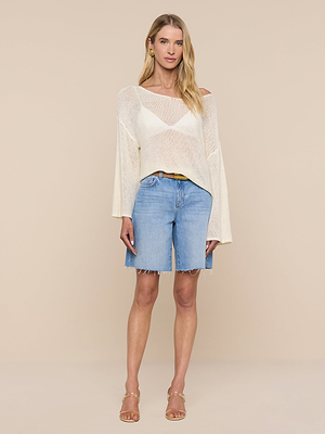 Woman stands against a beige background, wearing the Ethan Knit Pullover—a relaxed-fit sheer cream long-sleeve top—over a white bralette, light blue frayed denim knee-length shorts, and beige heeled sandals.