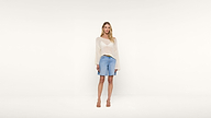 A woman stands against a plain white background, wearing a sheer long-sleeve sweater, denim knee-length shorts, and tan heeled sandals. She has long blonde hair and a relaxed posture.
