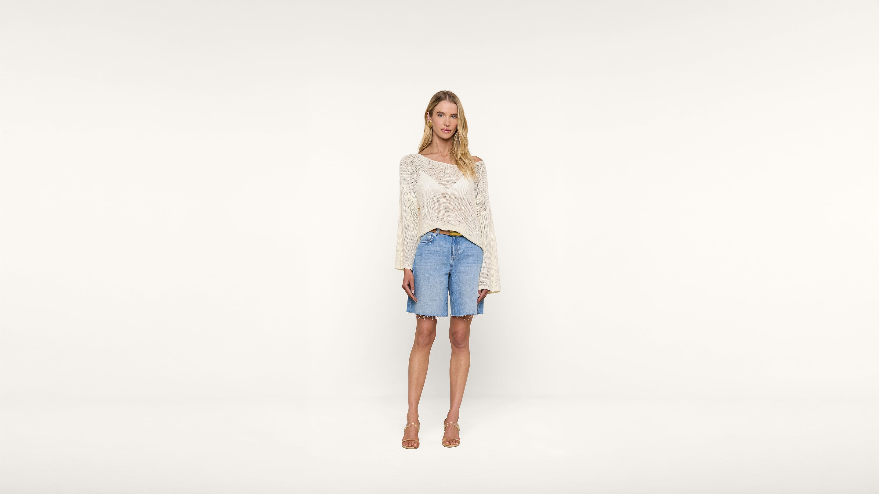A woman stands against a plain white background, wearing a sheer long-sleeve sweater, denim knee-length shorts, and tan heeled sandals. She has long blonde hair and a relaxed posture.