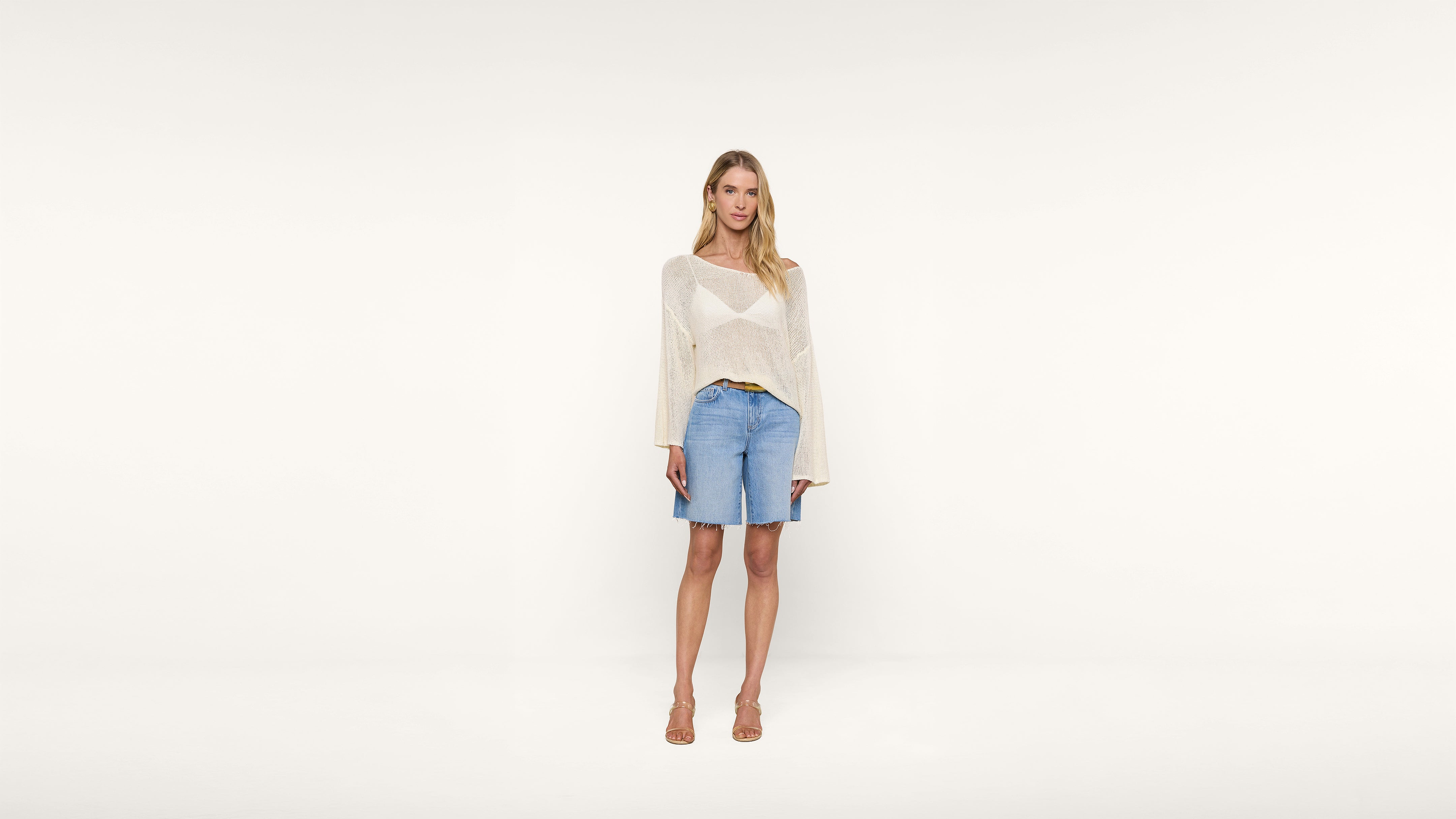 A woman stands against a plain white background, wearing a sheer long-sleeve sweater, denim knee-length shorts, and tan heeled sandals. She has long blonde hair and a relaxed posture.