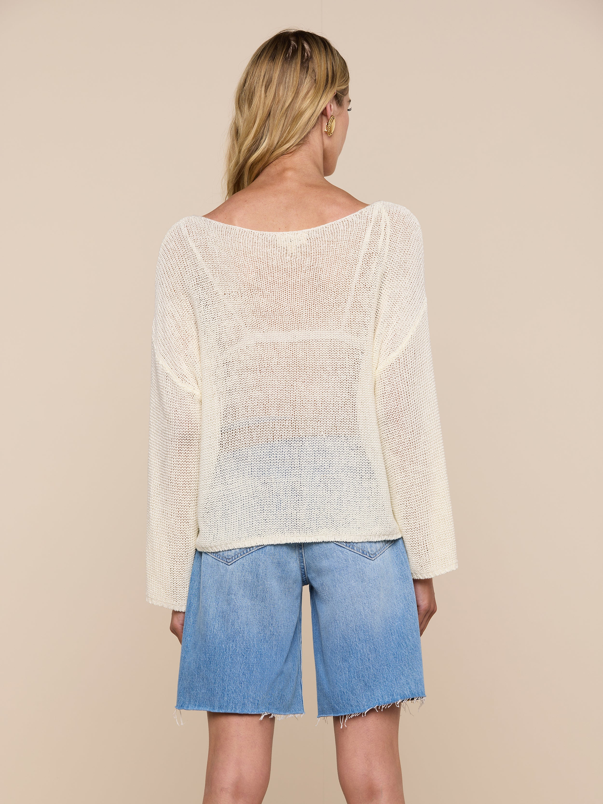 A woman with long blonde hair, seen from behind, wears the Ethan Knit Pullover with a relaxed fit and an off-shoulder neckline, paired with knee-length blue denim shorts against a plain light background.
